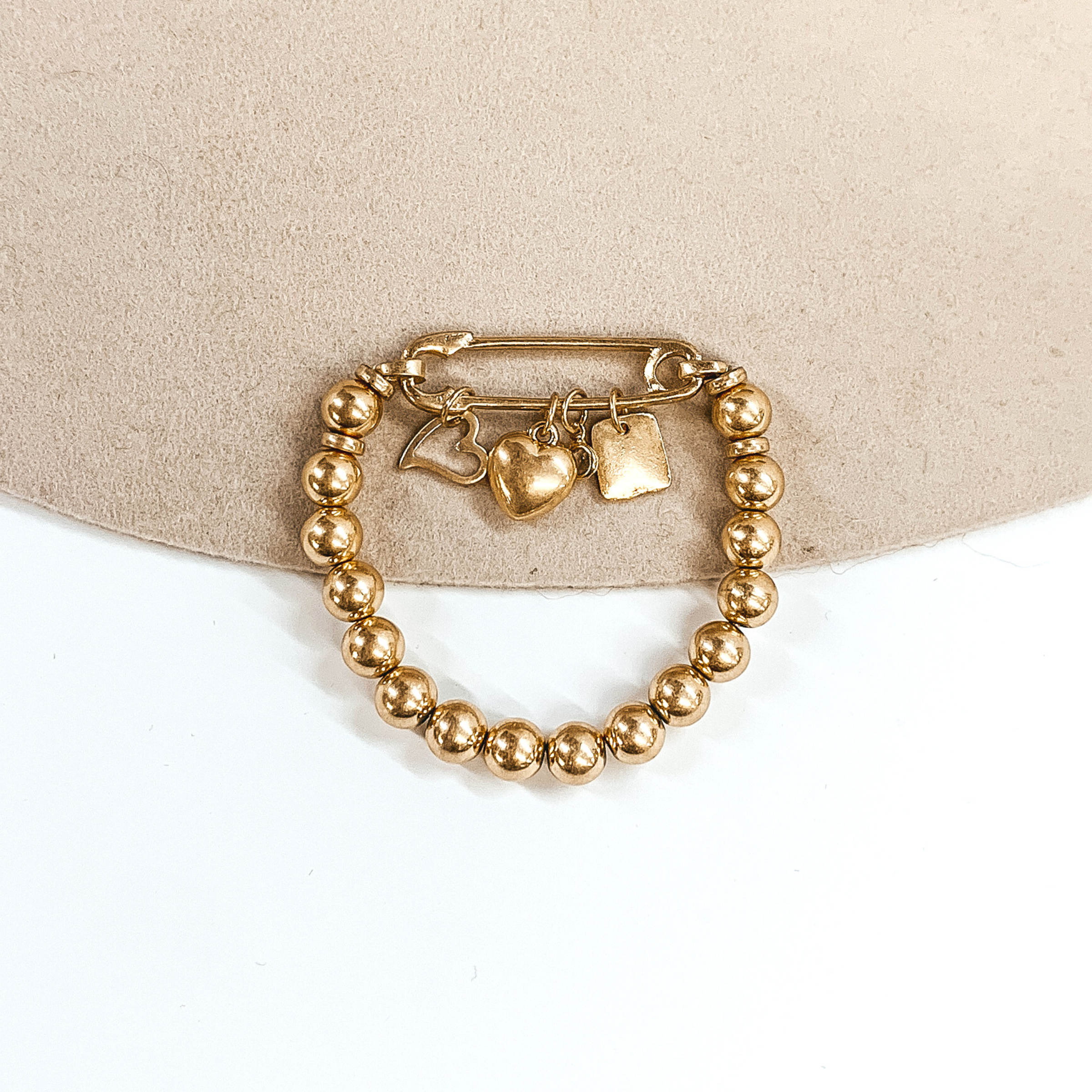 This is a gold beaded bracelet with a safety pin pendant closing the ends of the bracelet. Hanging on the gold safety pin are four gold charms including a square, a clear crystal, a heart, and an outlined heart. This bracelet is pictured on a beige and white background.