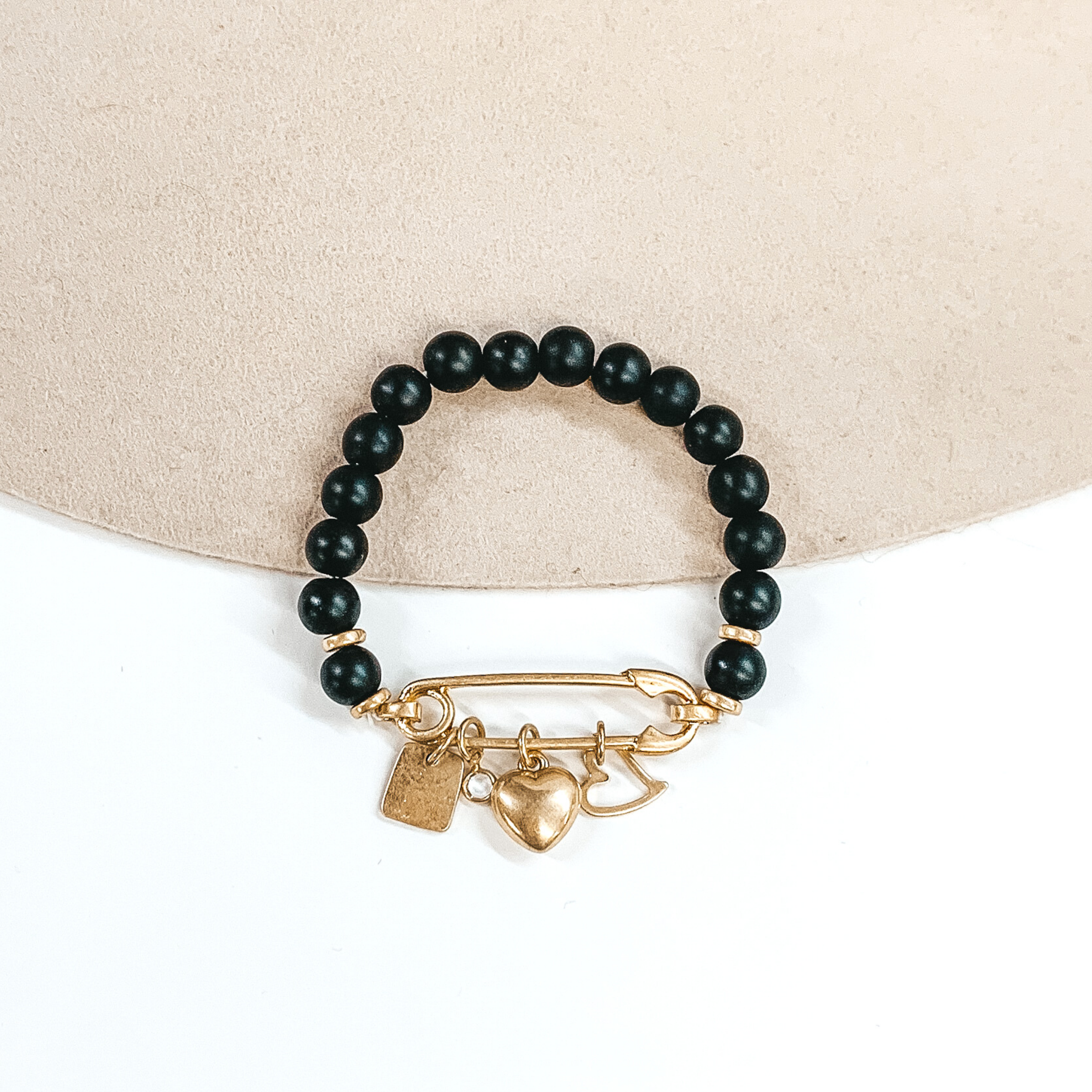 This is a black beaded bracelet with a safety pin pendant closing the ends of the bracelet. Hanging on the gold safety pin are four gold charms including a square, a clear crystal, a heart, and an outlined heart. This bracelet is pictured on a beige and white background.