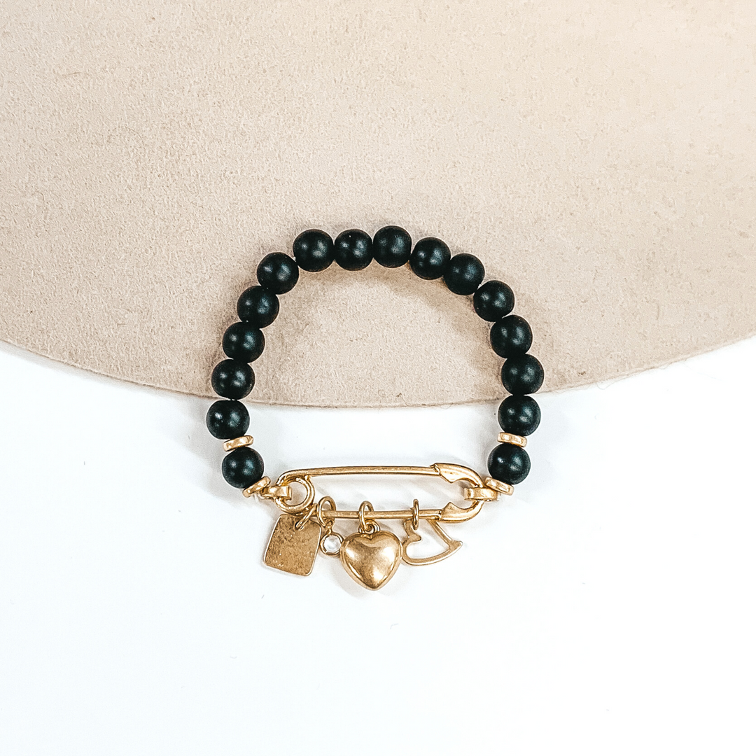 This is a black beaded bracelet with a safety pin pendant closing the ends of the bracelet. Hanging on the gold safety pin are four gold charms including a square, a clear crystal, a heart, and an outlined heart. This bracelet is pictured on a beige and white background.
