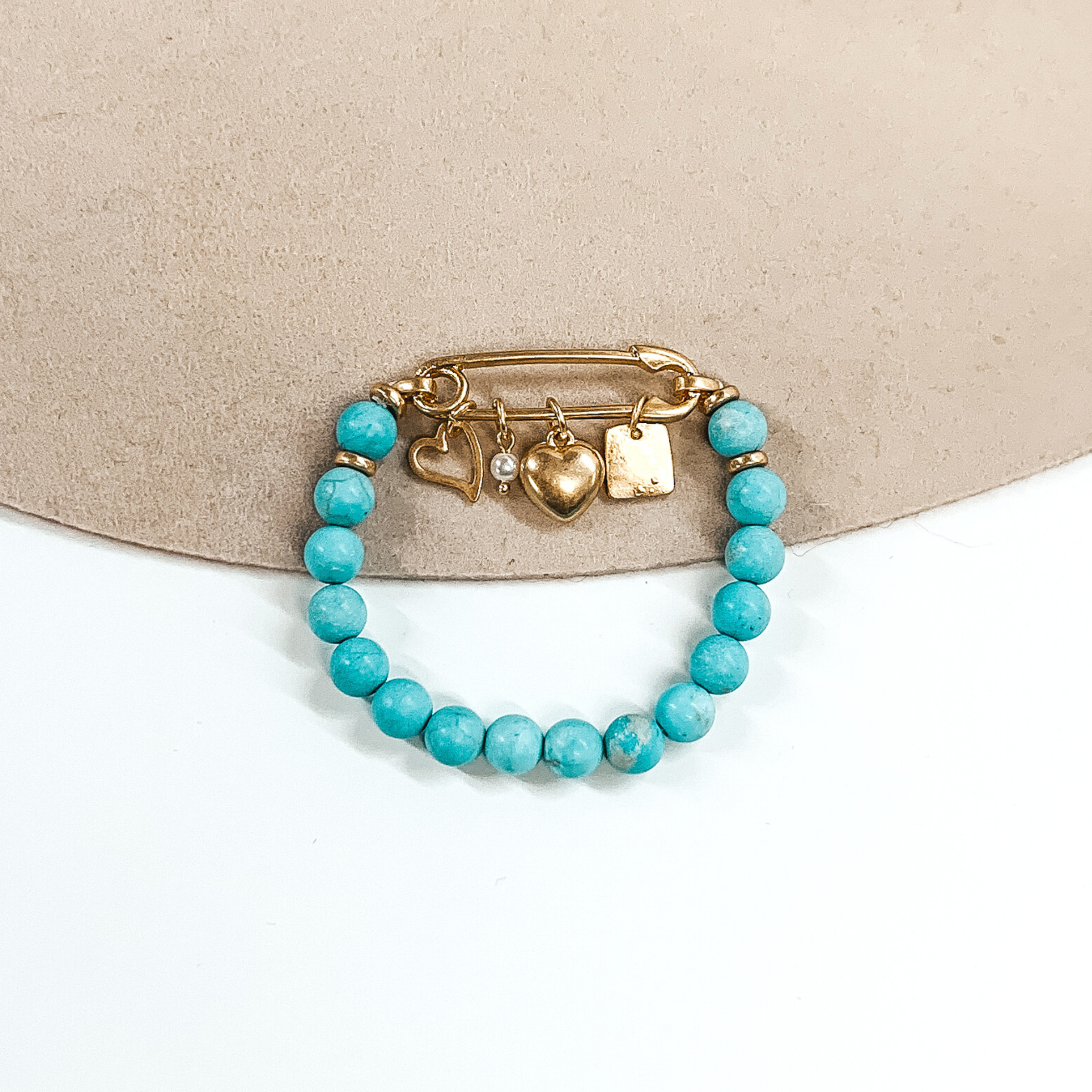 This is a turquoise beaded bracelet with a safety pin pendant closing the ends of the bracelet. Hanging on the gold safety pin are four gold charms including a square, a white pearl, a heart, and an outlined heart. This bracelet is pictured on a beige and white background.
