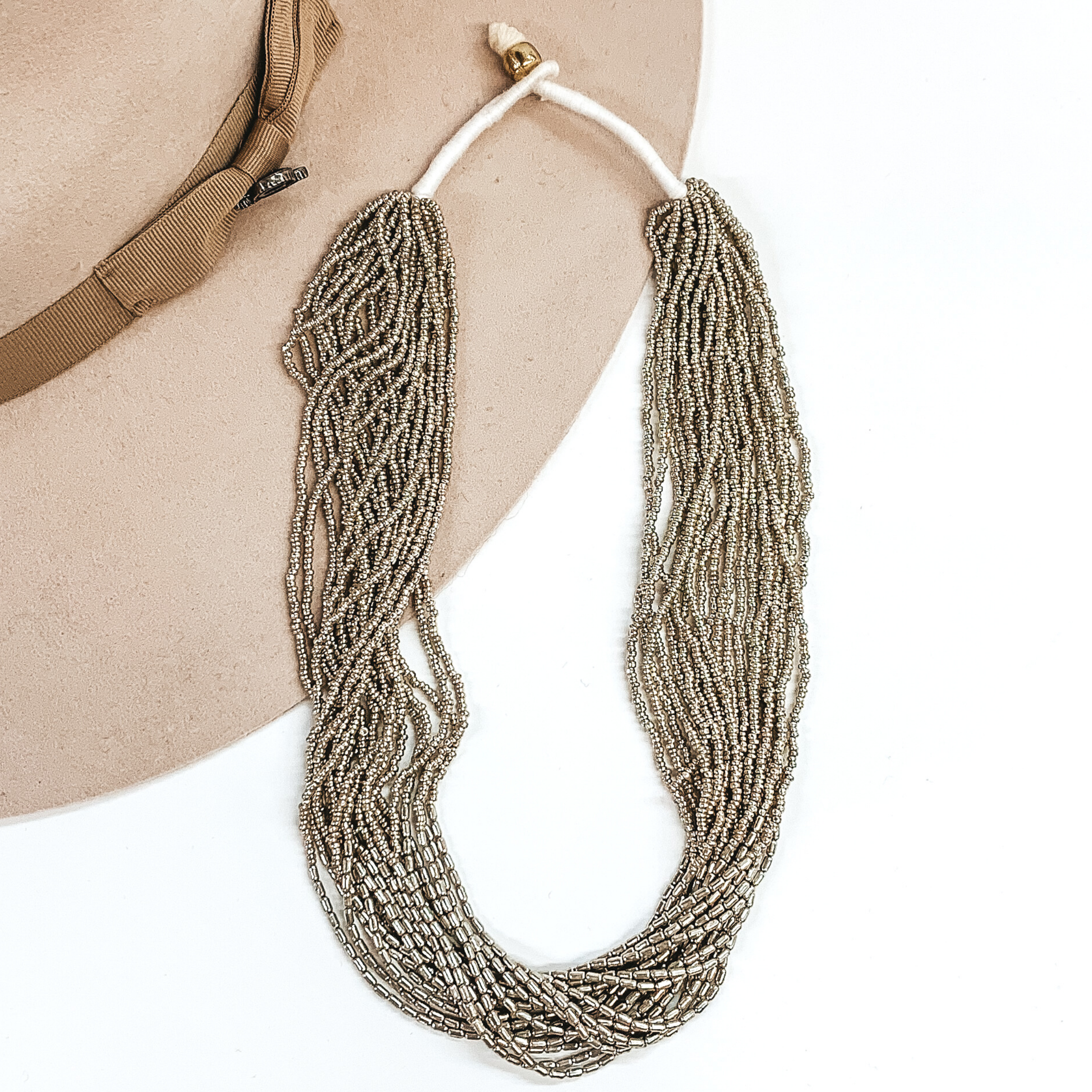 The strands on this necklace have silver beads and silver beads in the middle of the string. There are a lot of single strings put together to make a single necklace. The necklace has a gold bead that goes through a loop to put the necklace on. This necklace is pictured laying on a beige hat on a white background.