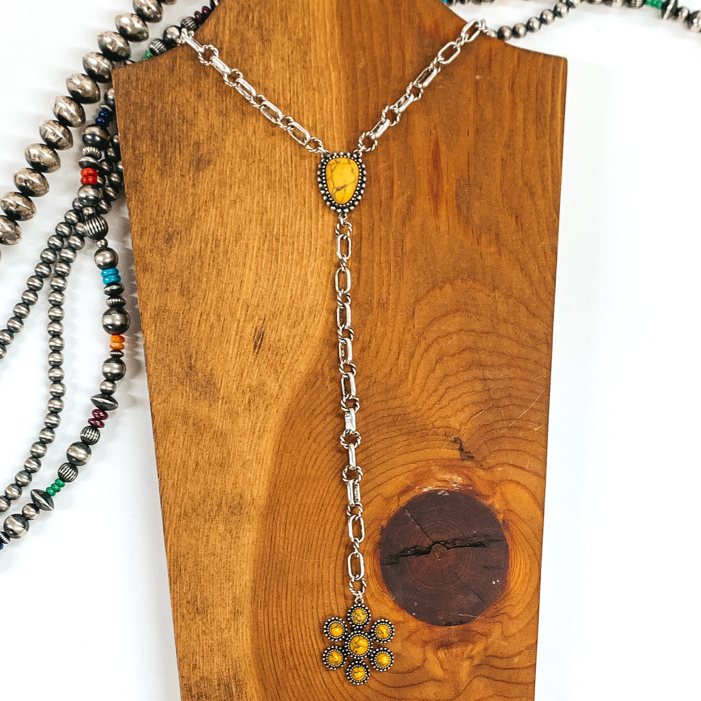 Silver chained necklace that is Navajo inspired with  an yellow irregular stoned pendant. Then hanging from the pendant, there is a hanging silver chain with a star burst pendant with yellow stones. This necklace is pictured on  wood necklace holder on a white background. It has different shaped and colored Navajo strands. 