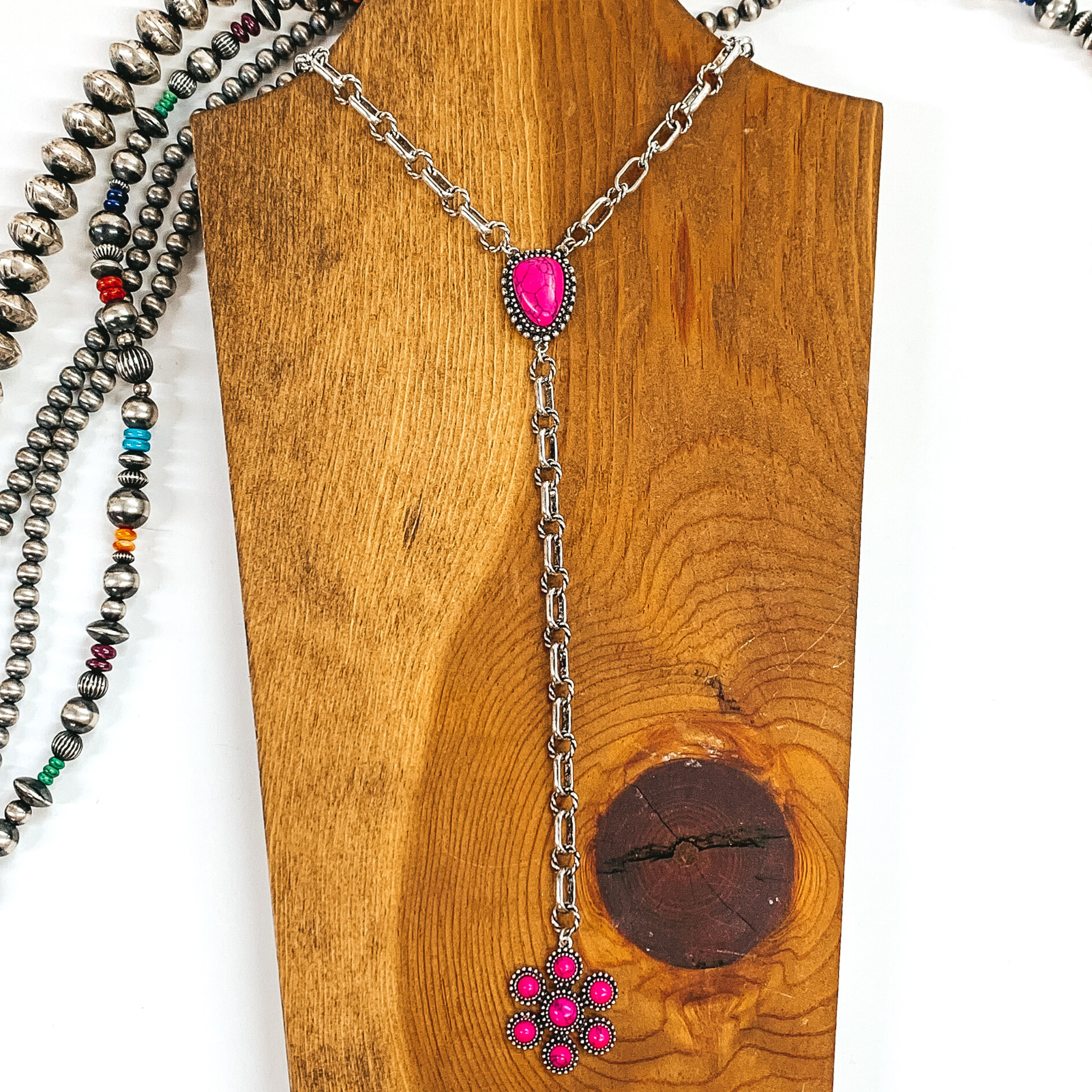 Silver chained necklace that is Navajo inspired with a pink irregular stoned pendant. Then hanging from the pendant, there is a hanging silver chain with a star burst pendant with pink stones. This necklace is pictured on  wood necklace holder on a white background. It has different shaped and colored Navajo strands. 
