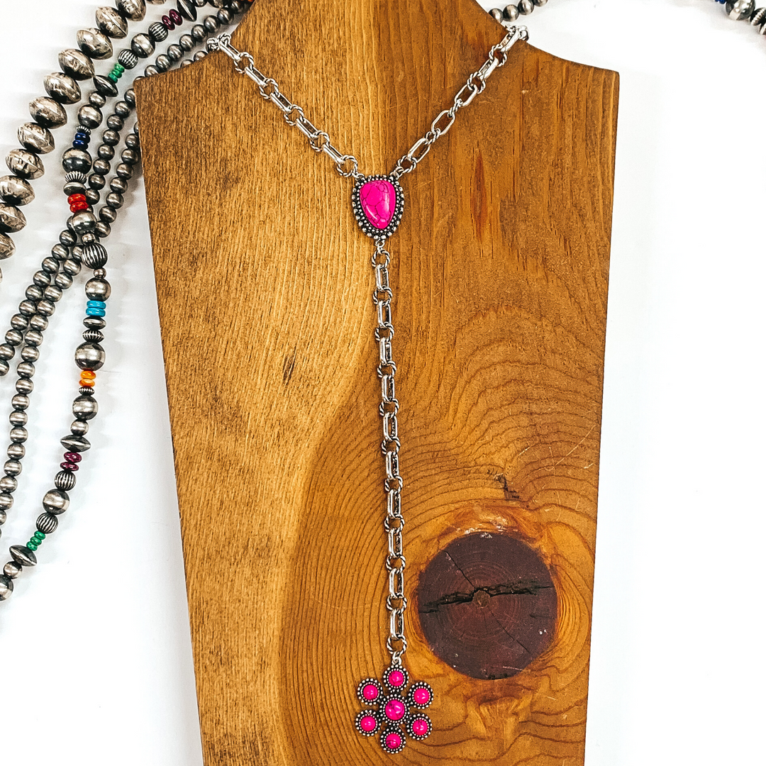 Silver chained necklace that is Navajo inspired with a pink irregular stoned pendant. Then hanging from the pendant, there is a hanging silver chain with a star burst pendant with pink stones. This necklace is pictured on  wood necklace holder on a white background. It has different shaped and colored Navajo strands. 