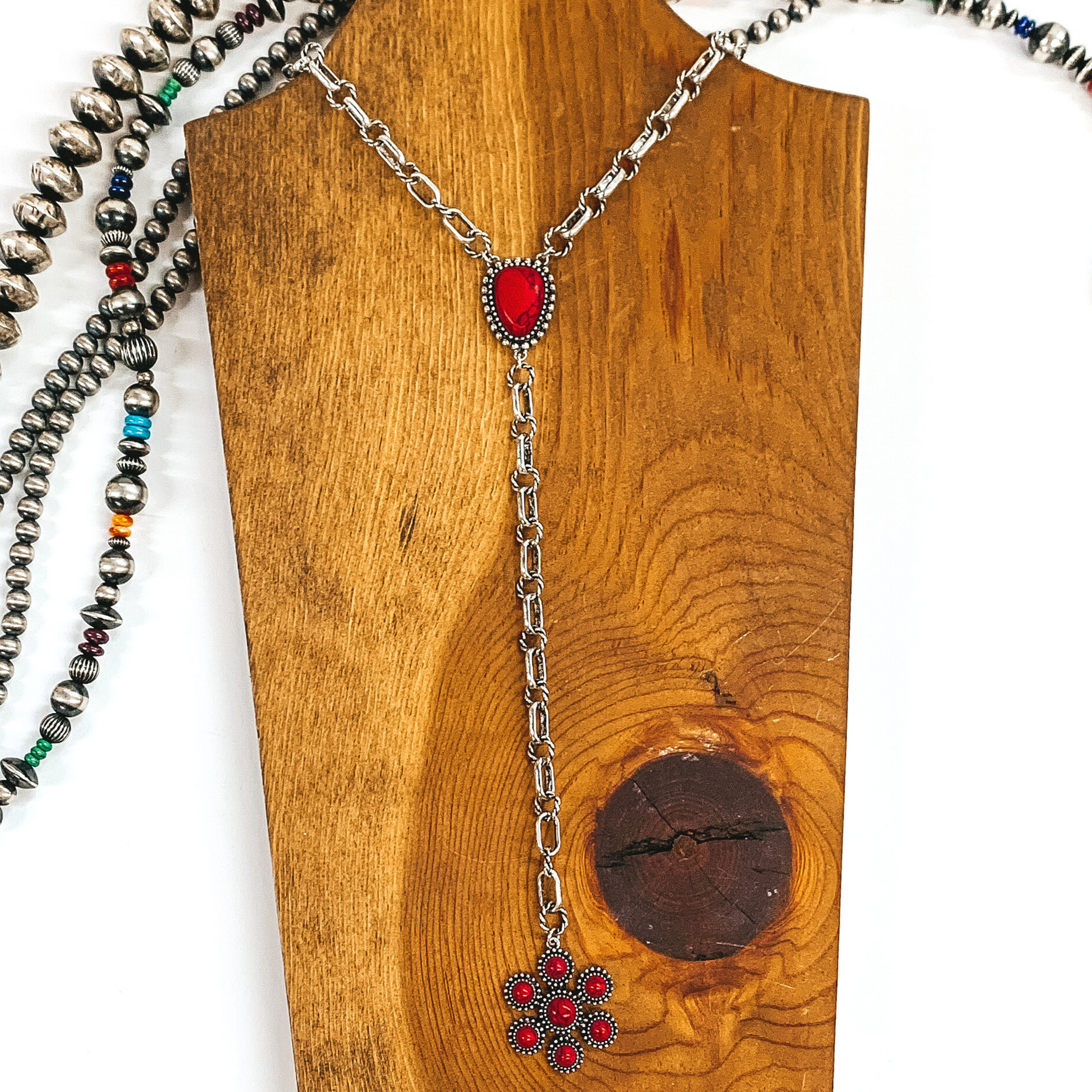 Silver chained necklace that is Navajo inspired with a red irregular stoned pendant. Then hanging from the pendant, there is a hanging silver chain with a star burst pendant with red stones. This necklace is pictured on  wood necklace holder on a white background. It has different shaped and colored Navajo strands. 