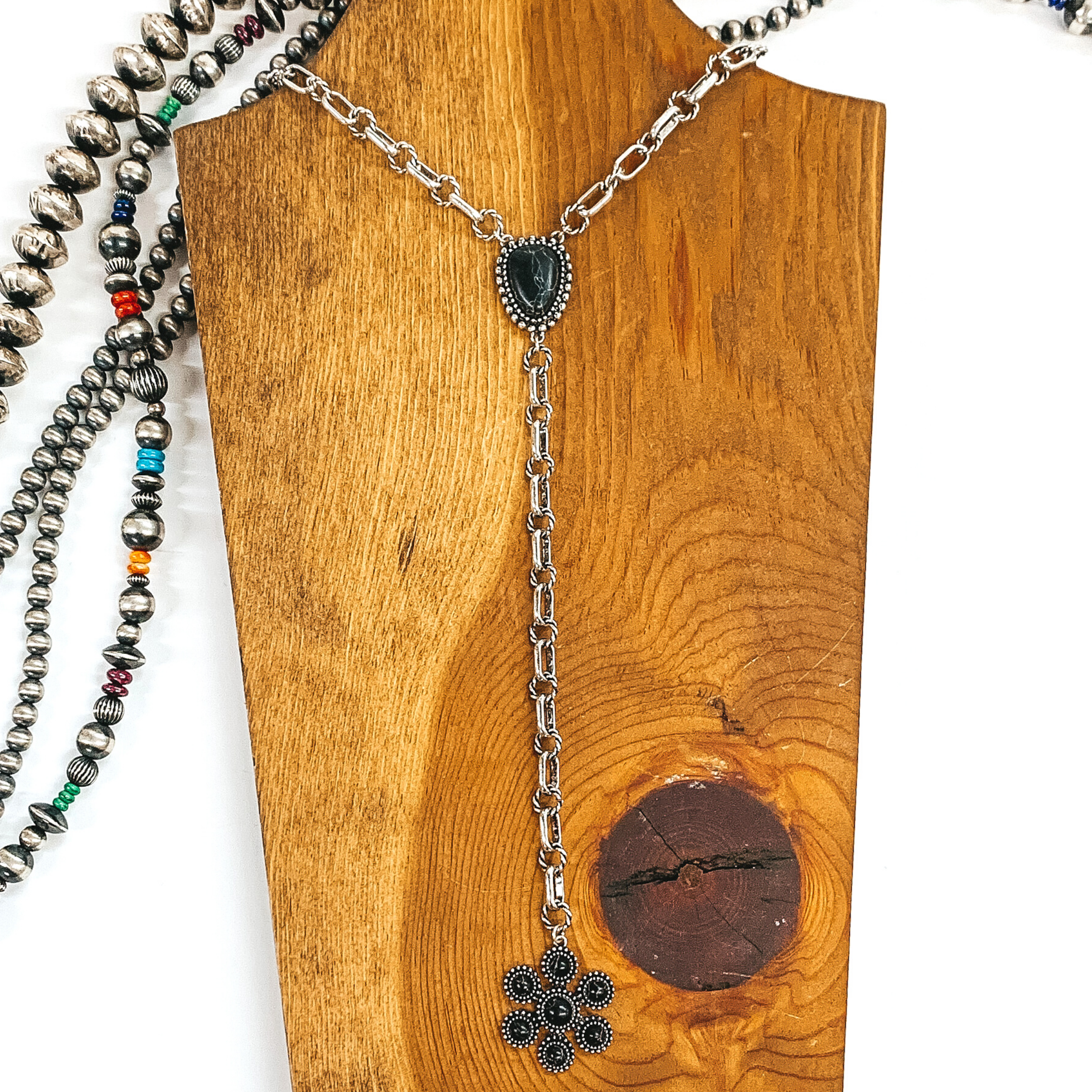 Silver chained necklace that is Navajo inspired with a black irregular stoned pendant. Then hanging from the pendant, there is a hanging silver chain with a star burst pendant with black stones. This necklace is pictured on  wood necklace holder on a white background. It has different shaped and colored Navajo strands. 