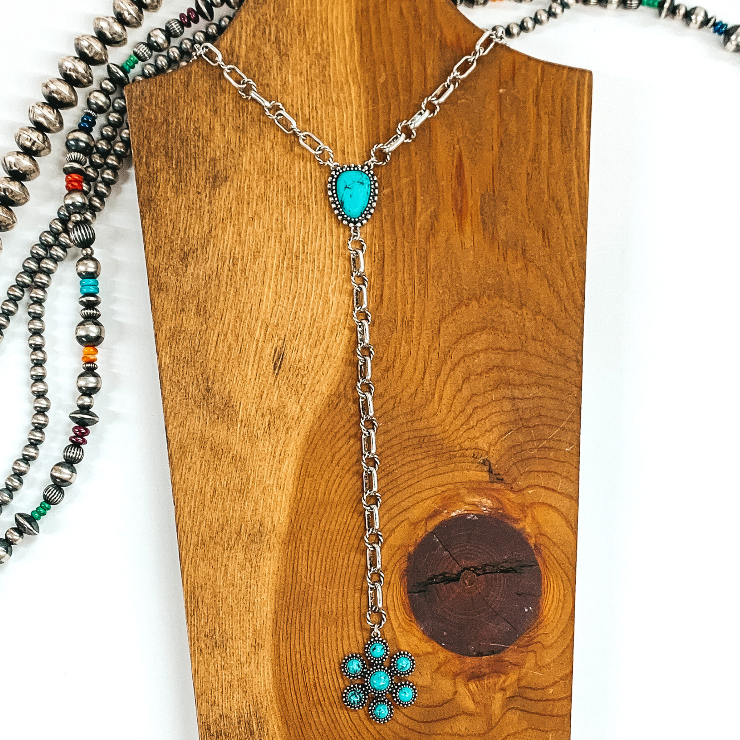 Silver chained necklace that is Navajo inspired with a turquoise irregular stoned pendant. Then hanging from the pendant, there is a hanging silver chain with a star burst pendant with turquoise stones. This necklace is pictured on  wood necklace holder on a white background. It has different shaped and colored Navajo strands. 