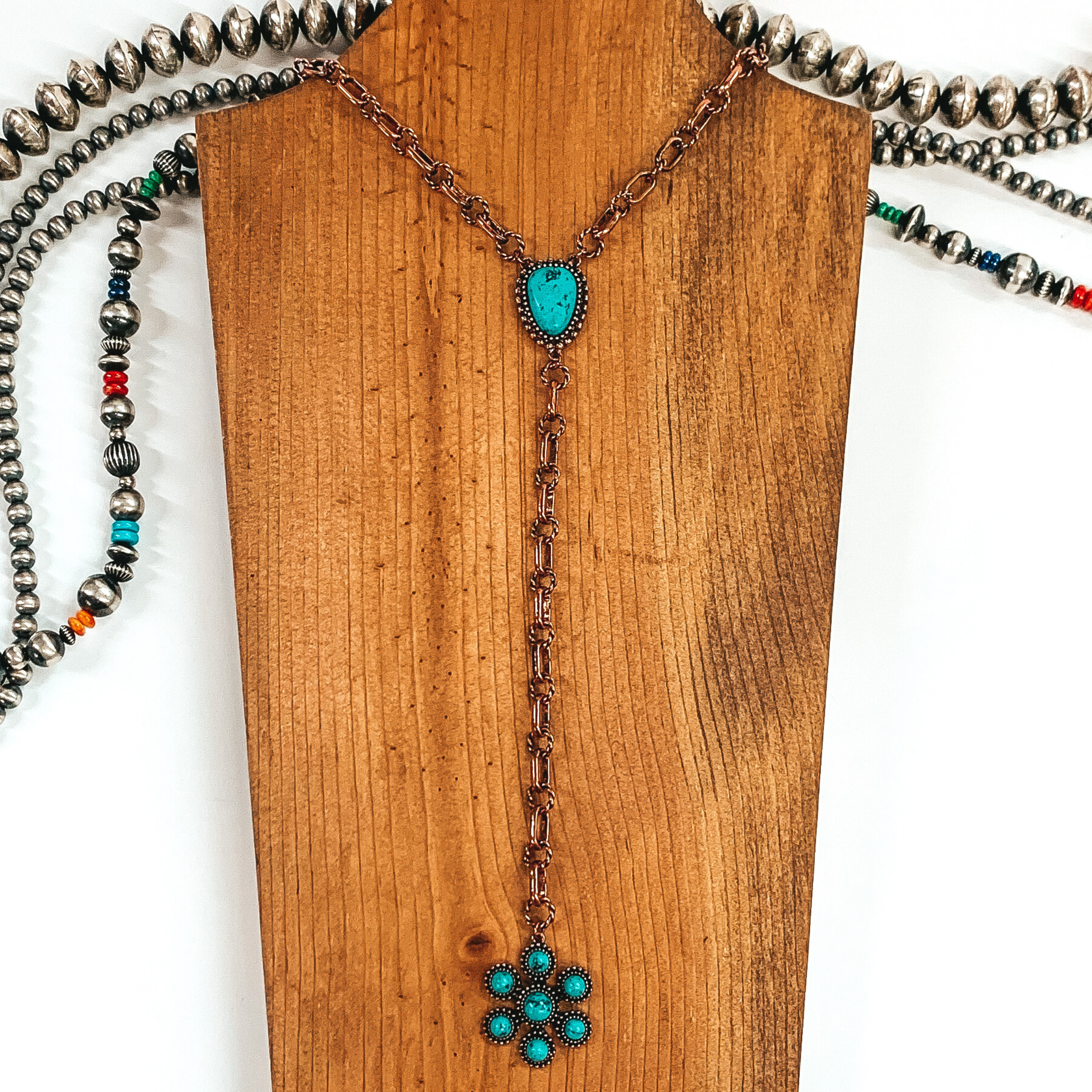 Copper chained necklace that is Navajo inspired with a turquoise irregular stoned pendant. Then hanging from the pendant, there is a hanging copper chain with a star burst pendant with turquoise stones. This necklace is pictured on  wood necklace holder on a white background. It has different shaped and colored Navajo strands. 