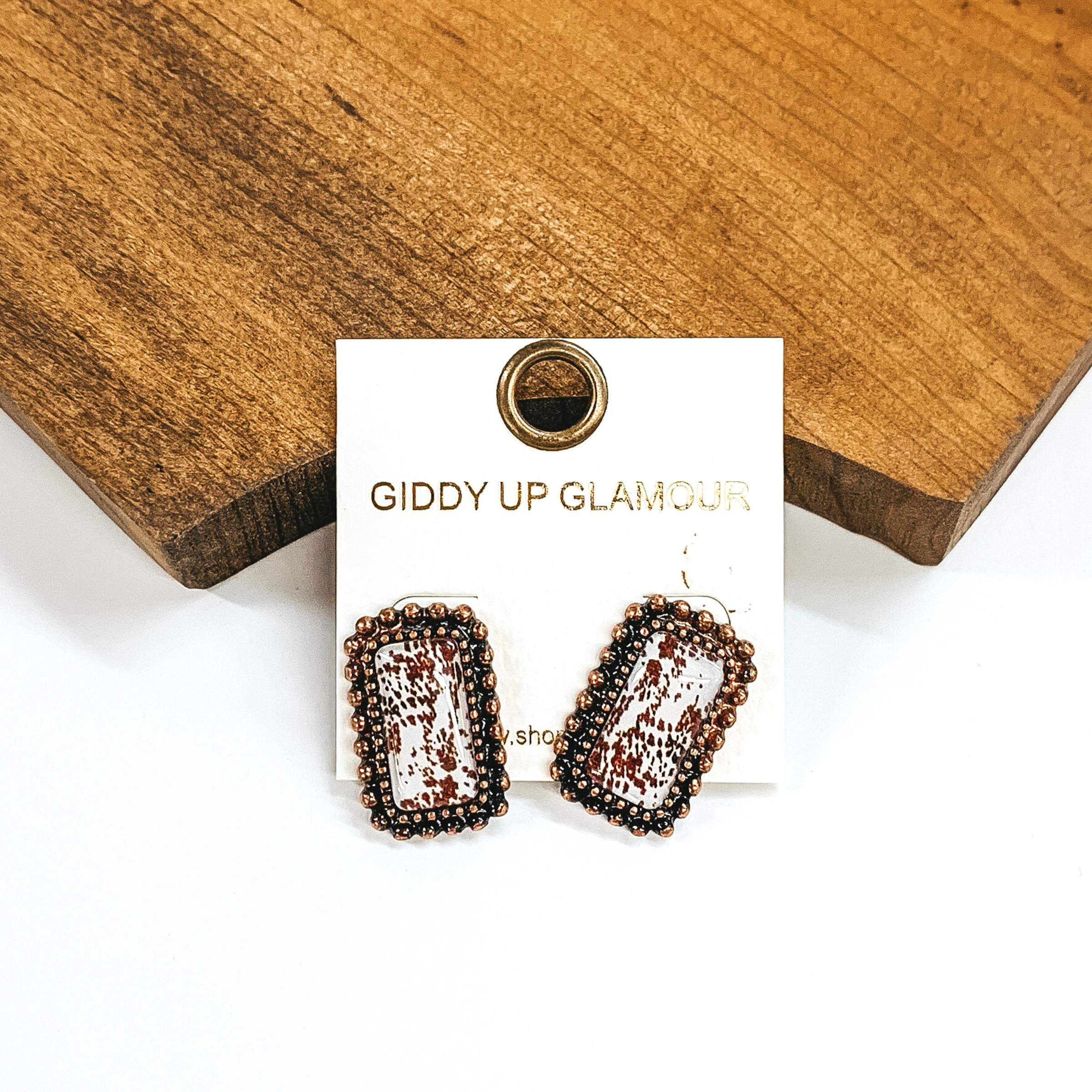 Rectangle stud earrings with brown cow print inlay. These earrings are outline with two rows of silver beads. These earrings are pictured laying against a block of wood on a white background. 