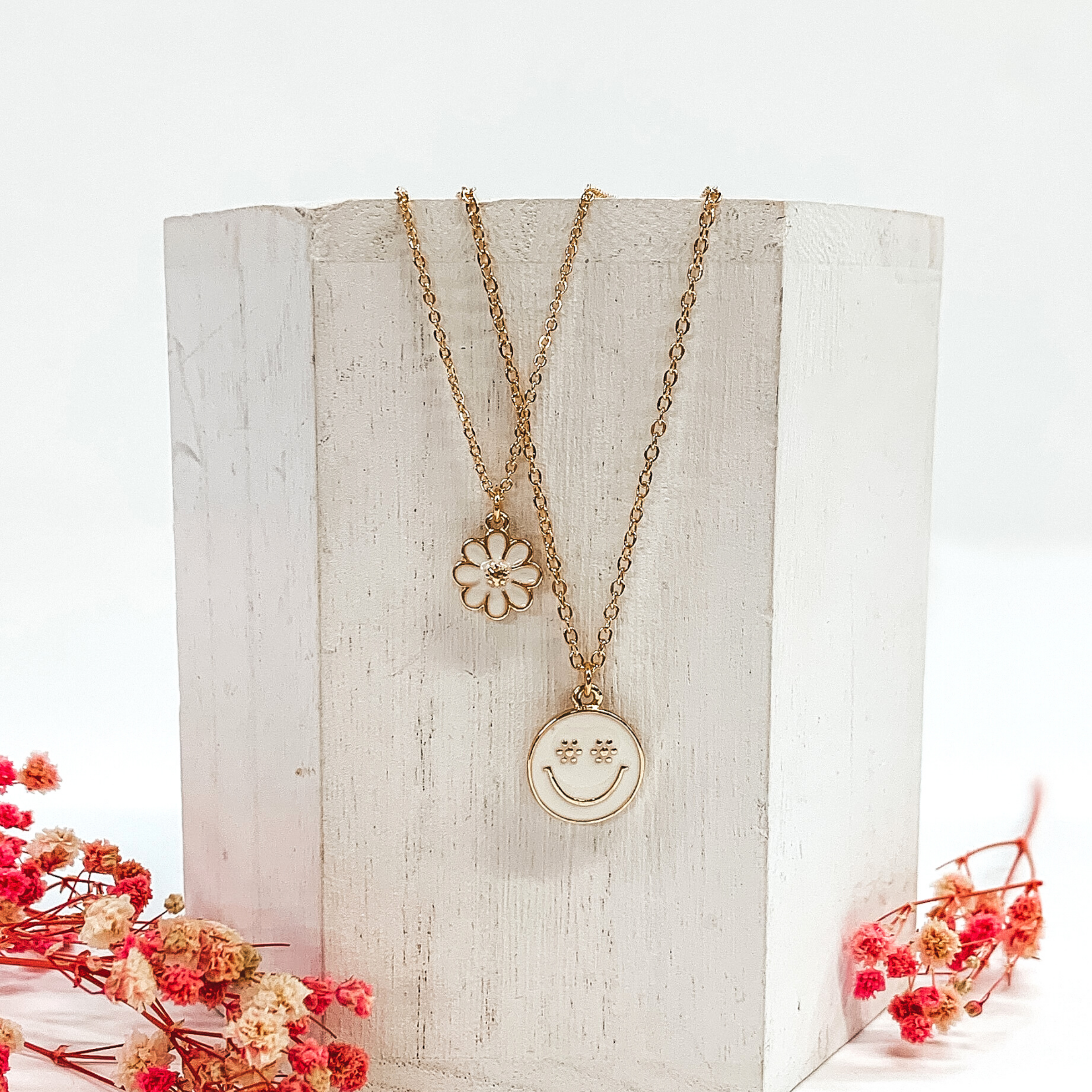 Gold double chained necklace. One strand has a white flower pendant and the other strand is longer and has a white circle pendant with a gold smiley face. This necklace is pictured laying on a white block on a white background with pink baby breath flowers at the bottom of the picture. 