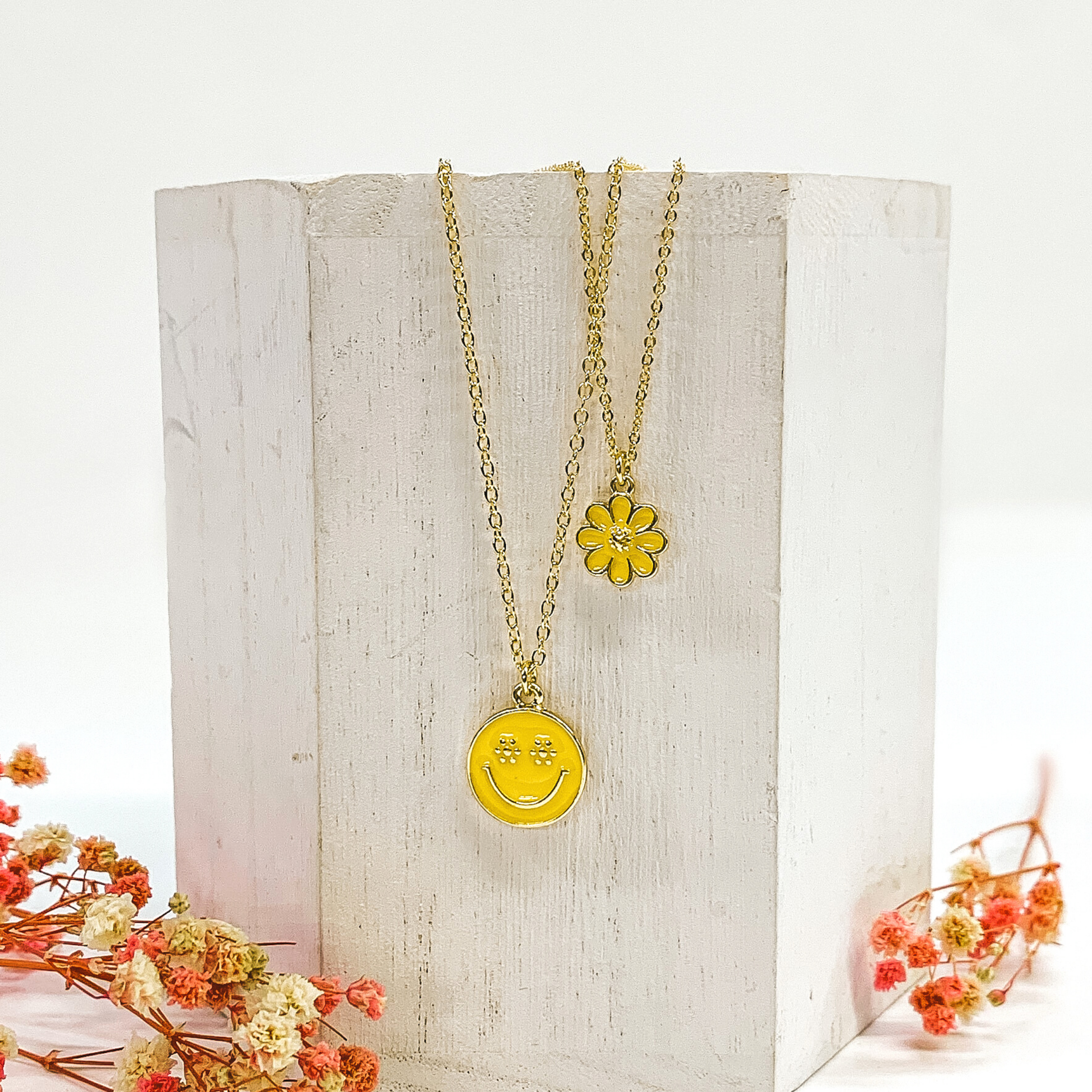 Gold double chained necklace. One strand has a yellow flower pendant and the other strand is longer and has a yellow circle pendant with a gold smiley face. This necklace is pictured laying on a white block on a white background with pink baby breath flowers at the bottom of the picture. 