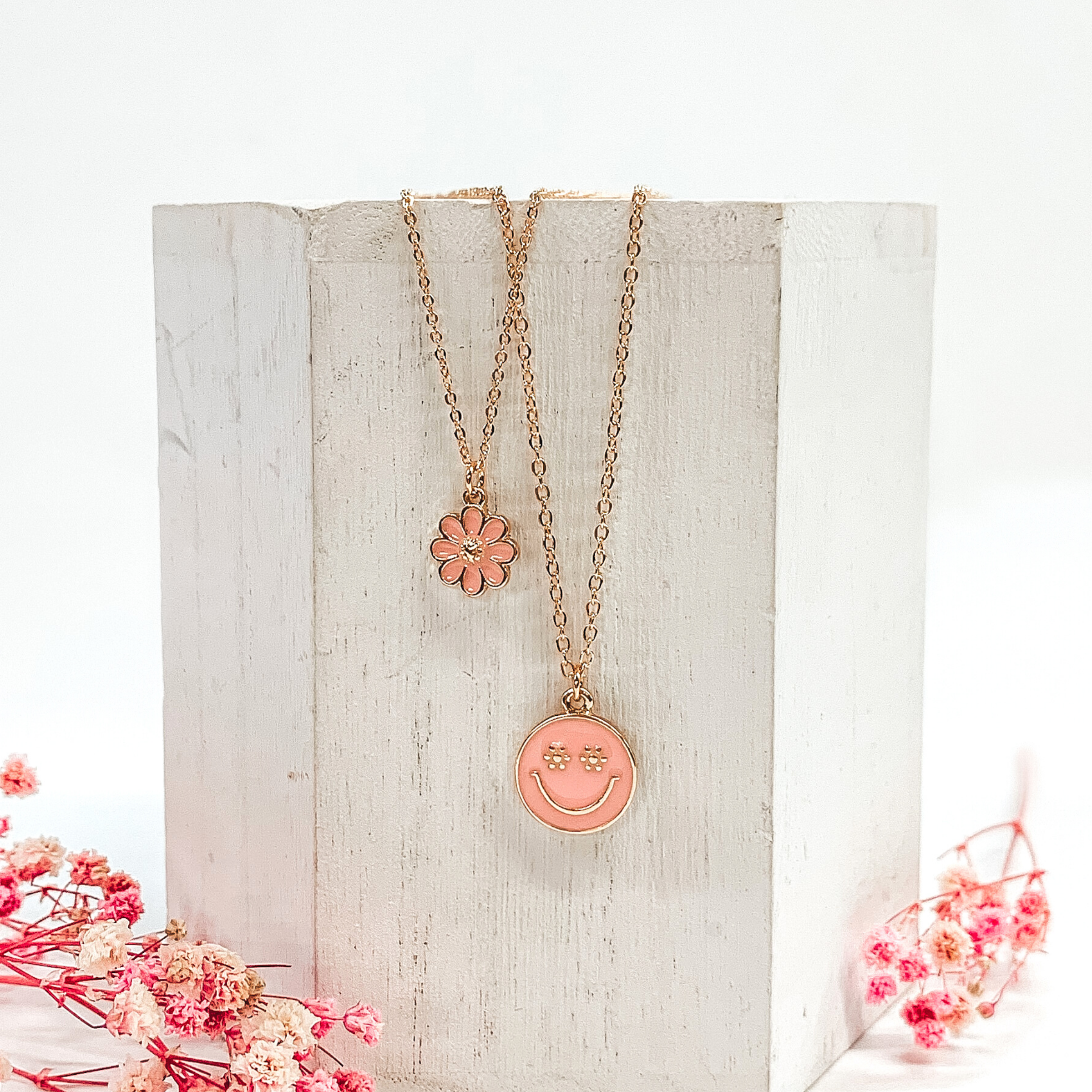 Gold double chained necklace. One strand has a baby pink flower pendant and the other strand is longer and has a baby pink circle pendant with a gold smiley face. This necklace is pictured laying on a white block on a white background with pink baby breath flowers at the bottom of the picture. 