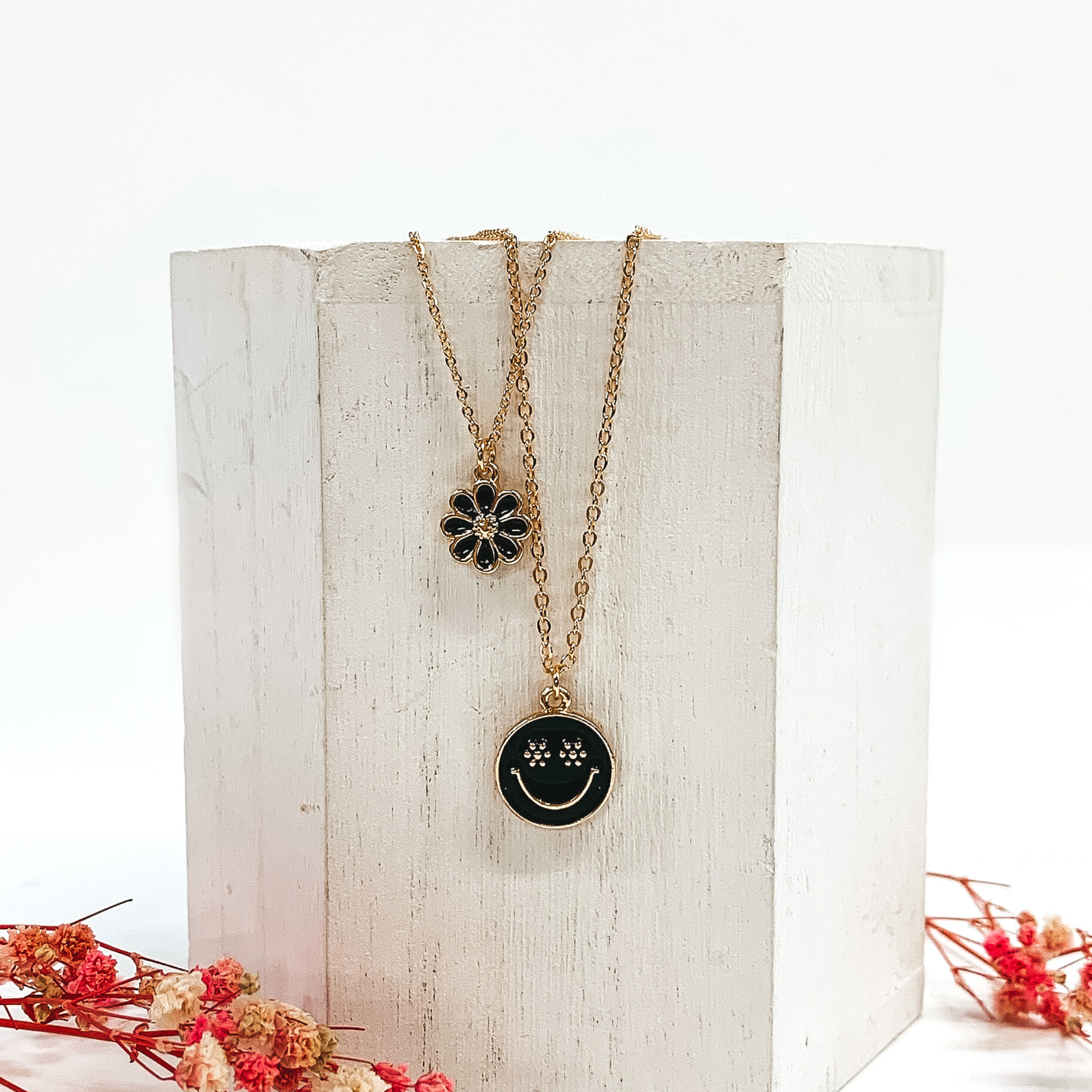 Gold double chained necklace. One strand has a black flower pendant and the other strand is longer and has a black circle pendant with a gold smiley face. This necklace is pictured laying on a white block on a white background with pink baby breath flowers at the bottom of the picture.