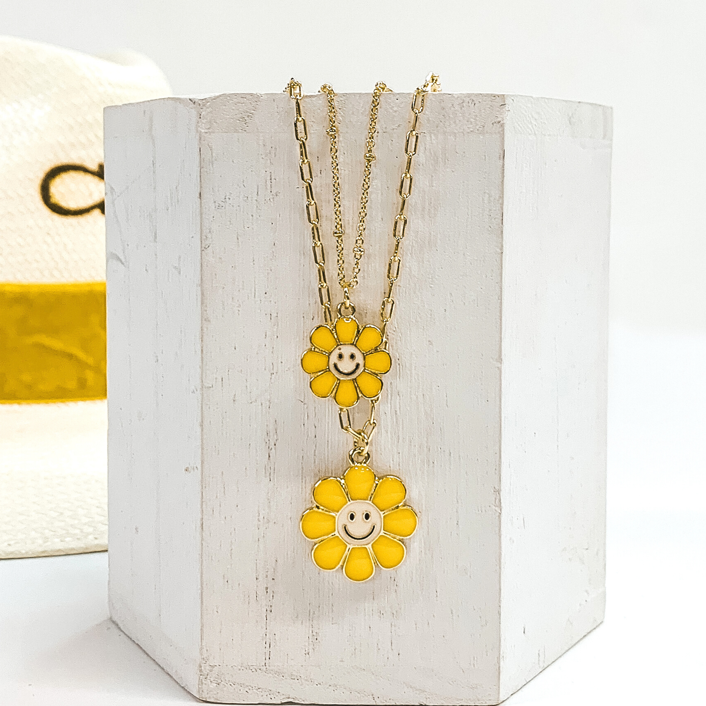 Gold double chained necklace. One strand has a yellow flower pendant with a white center and black smiley face. The other strand is longer and has a slightly bigger yellow flower pendant with a white center and black smiley face. This necklace is pictured laying on a white block on a white background with a straw hat with a yellow band in the background. 