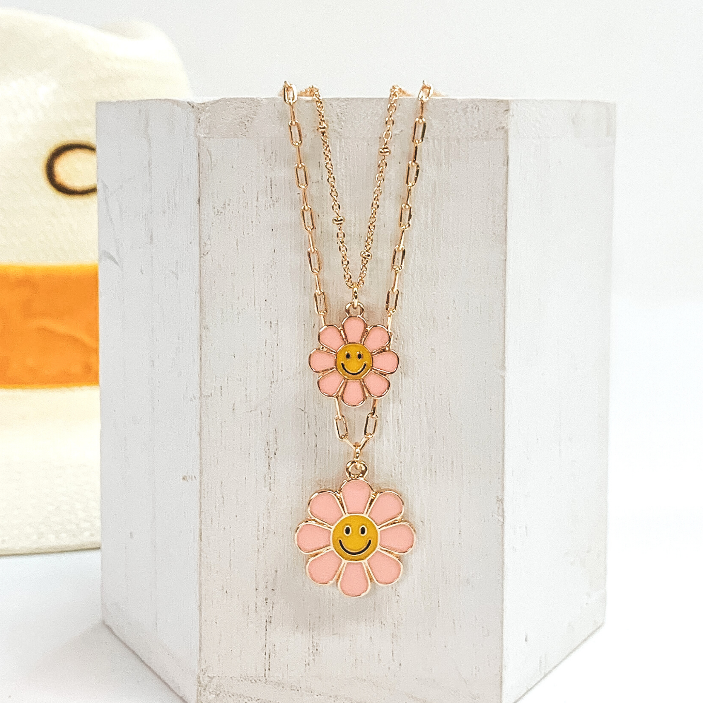 Gold double chained necklace. One strand has a baby pink flower pendant with a yellow center and black smiley face. The other strand is longer and has a slightly bigger baby pink flower pendant with a yellow center and black smiley face. This necklace is pictured laying on a white block on a white background with a straw hat with a yellow band in the background. 