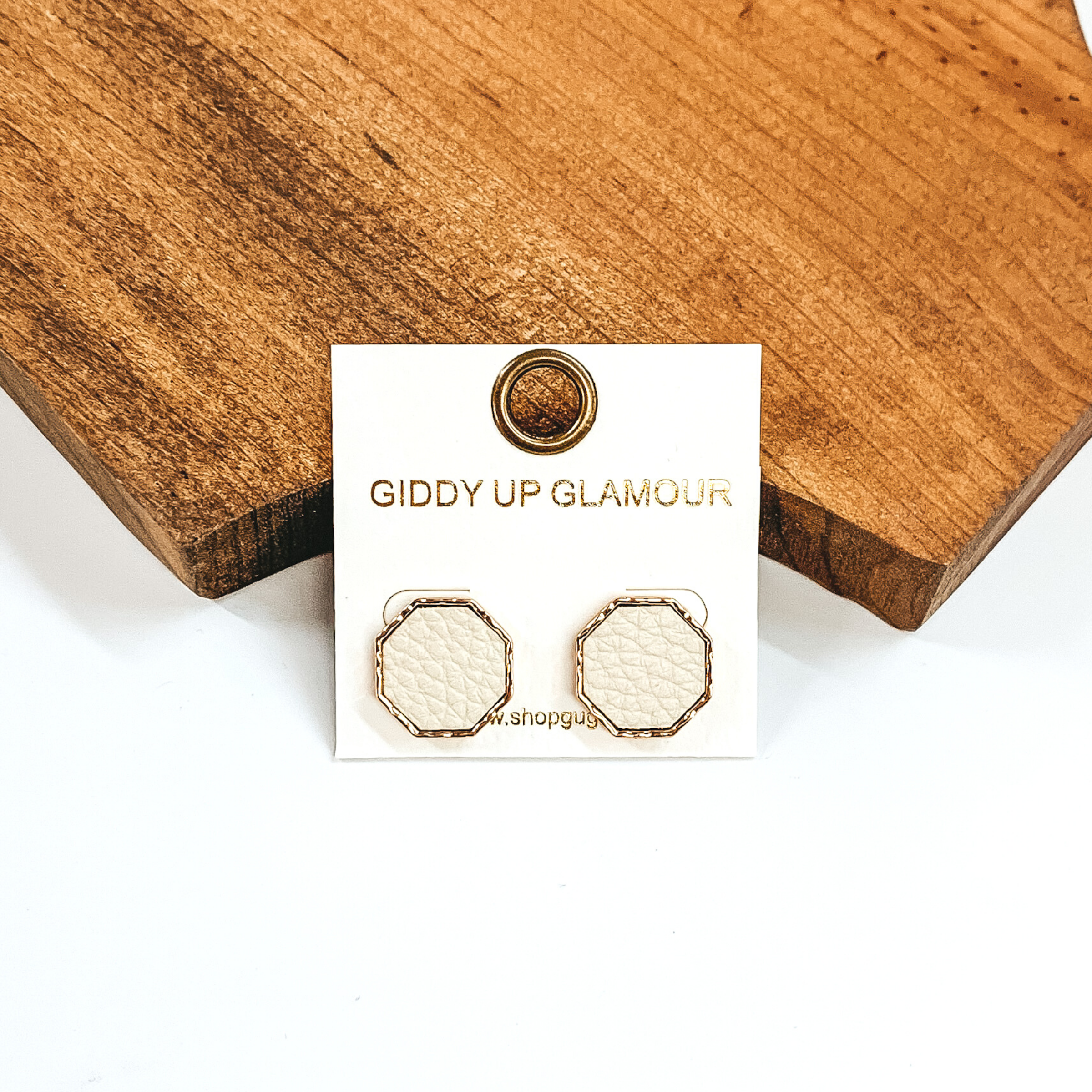 Gold, hexagon stud earrings with an ivory leather like inlay. Pictured on a white background in front of a piece of wood. 