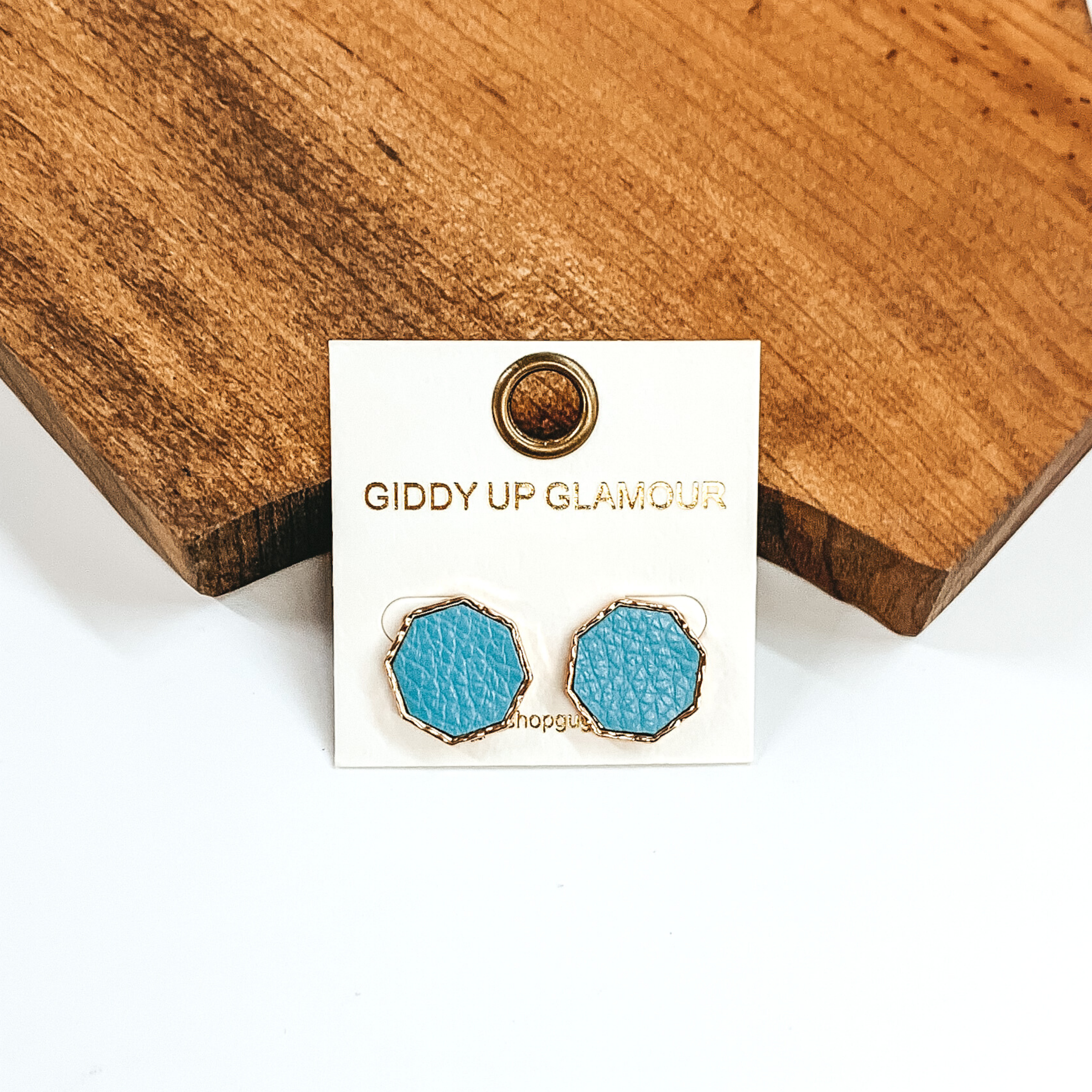 Gold, hexagon stud earrings with an pale blue leather like inlay. Pictured on a white background in front of a piece of wood.