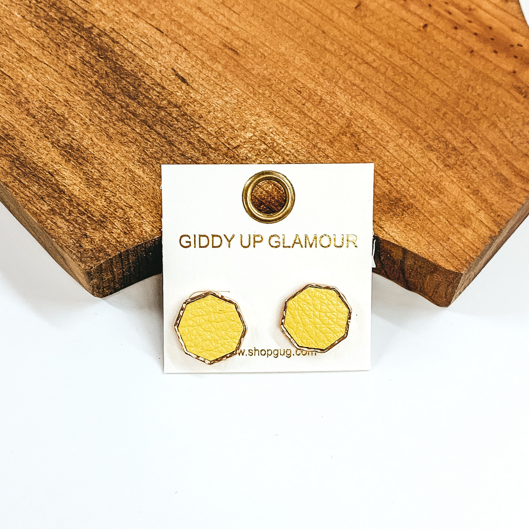 Gold, hexagon stud earrings with a pale yellow leather like inlay. Pictured on a white background in front of a piece of wood. 