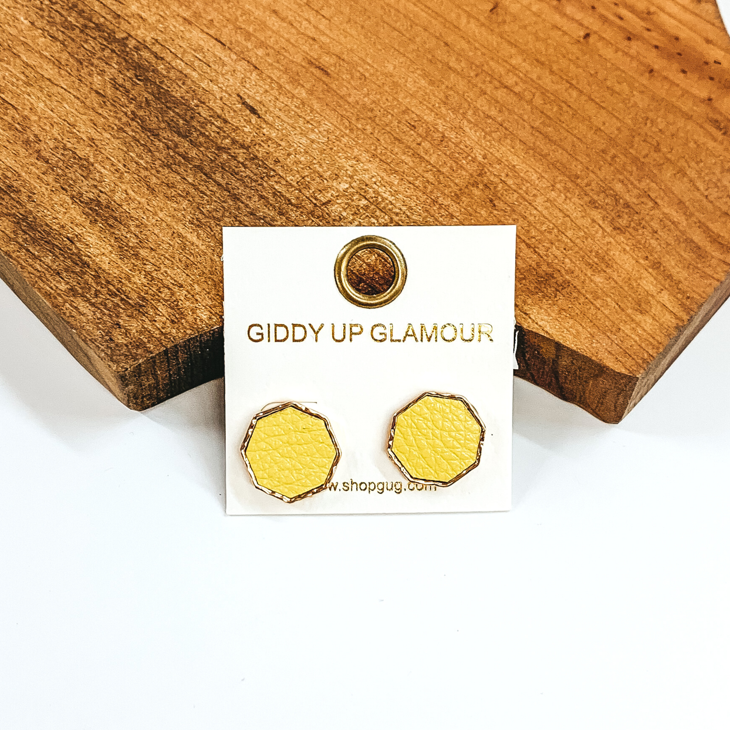 Gold, hexagon stud earrings with a pale yellow leather like inlay. Pictured on a white background in front of a piece of wood. 