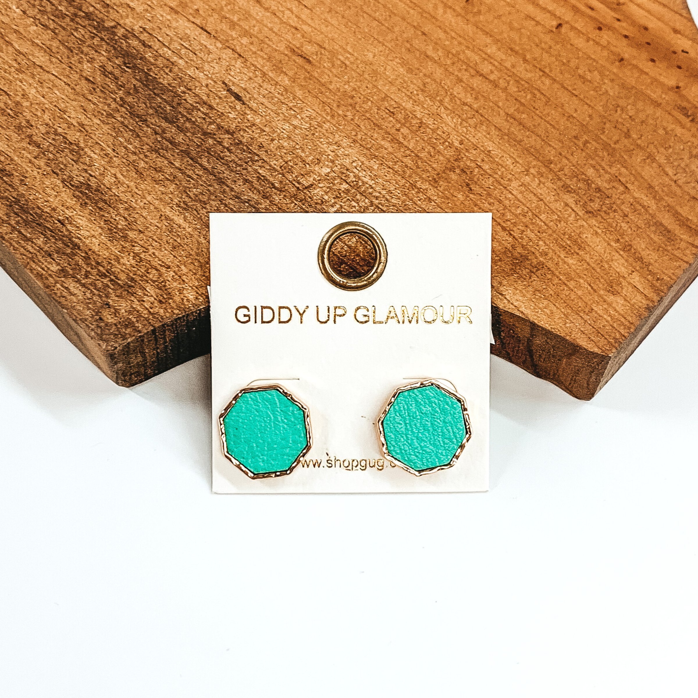Gold, hexagon stud earrings with a green leather like inlay. Pictured on a white background in front of a piece of wood. 