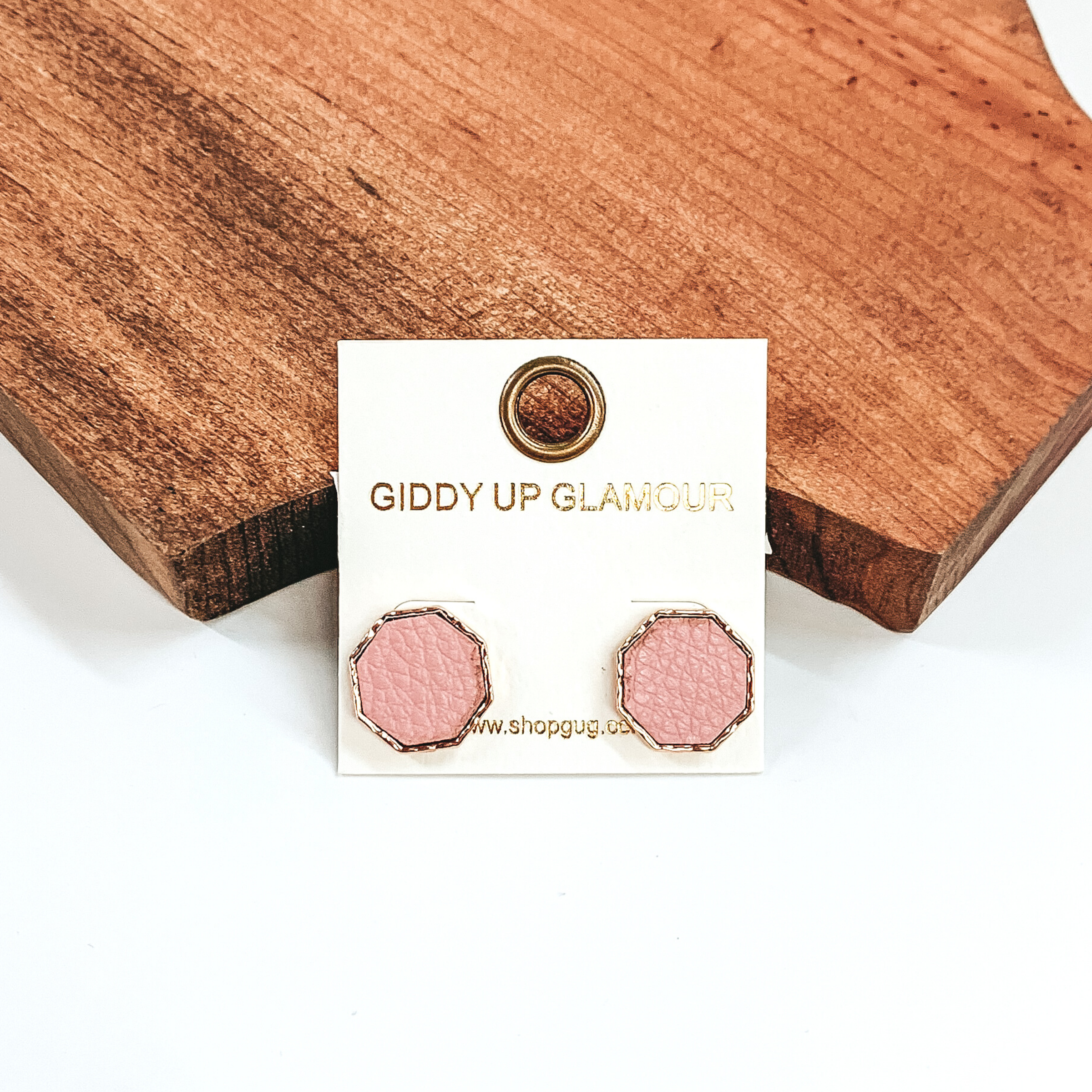 Gold, hexagon stud earrings with a pale pink leather like inlay. Pictured on a white background in front of a piece of wood. 