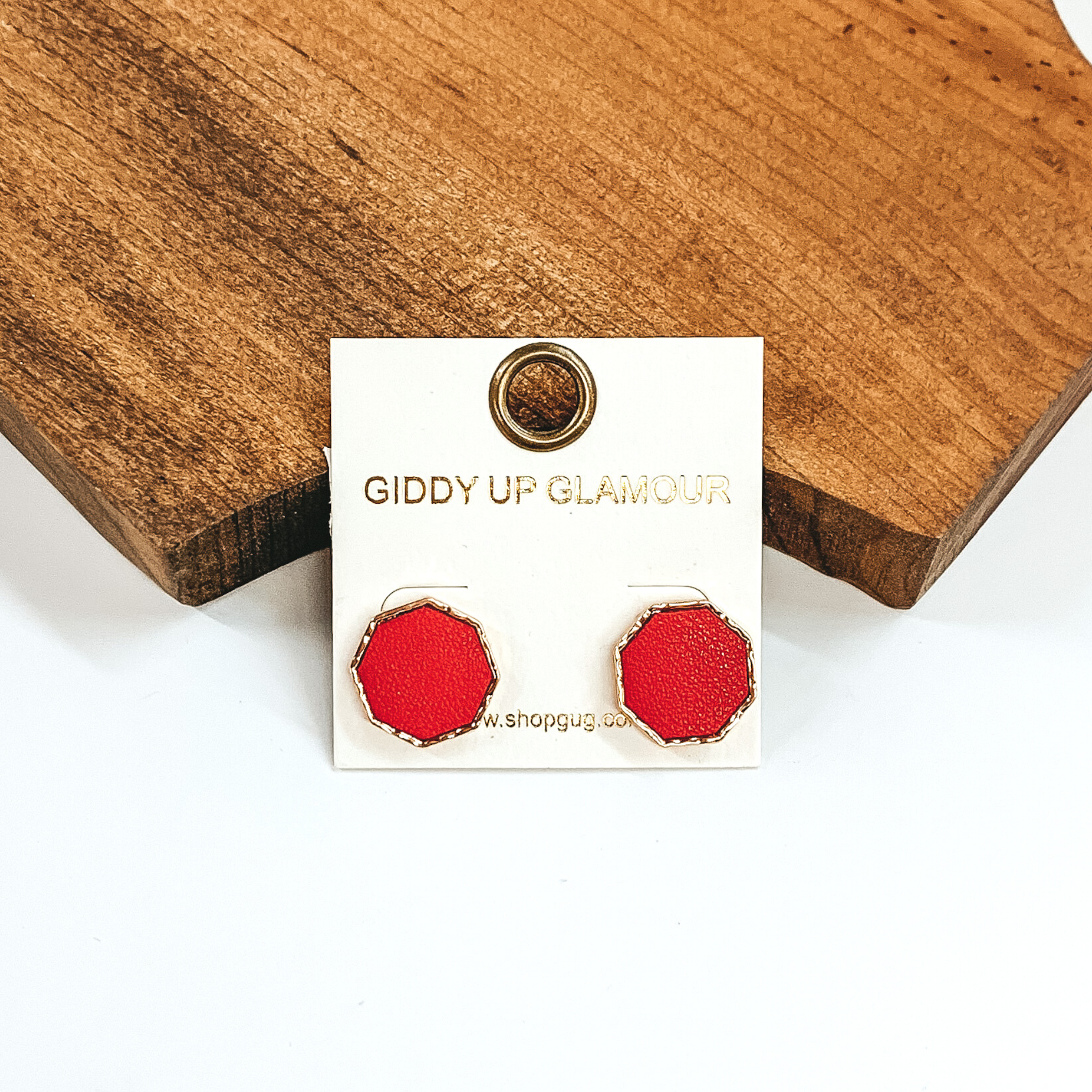 Gold, hexagon stud earrings with a red leather like inlay. Pictured on a white background in front of a piece of wood. 