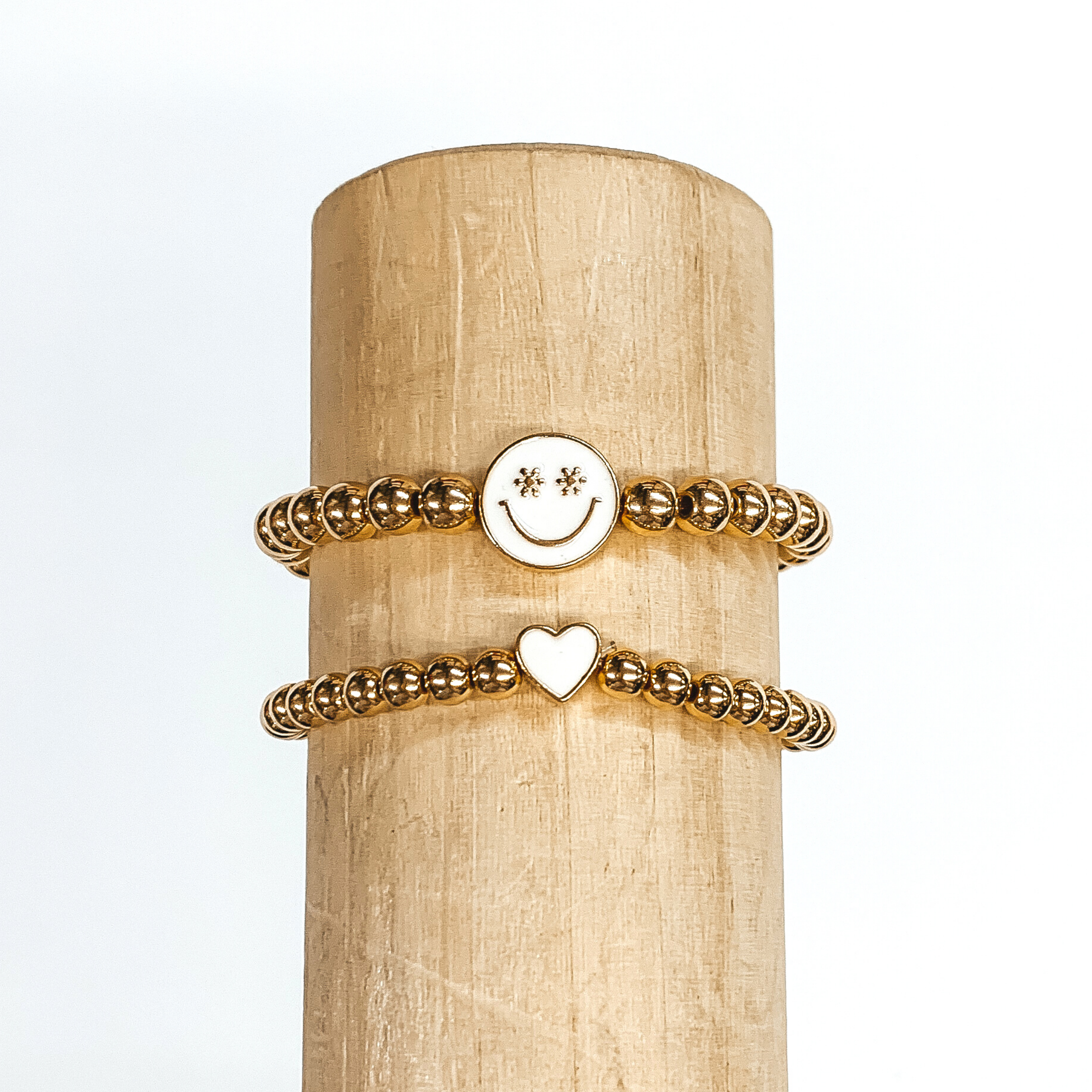 Two gold beaded bracelets. One bracelet has a tiny heart charm colored ivory. The other bracelet has a circle charm that is colored ivory with a smiley face in gold in the center. The eyes of the smiley face are tiny flowers. These two bracelets are pictured on a wood bracelet holder on a white background. 