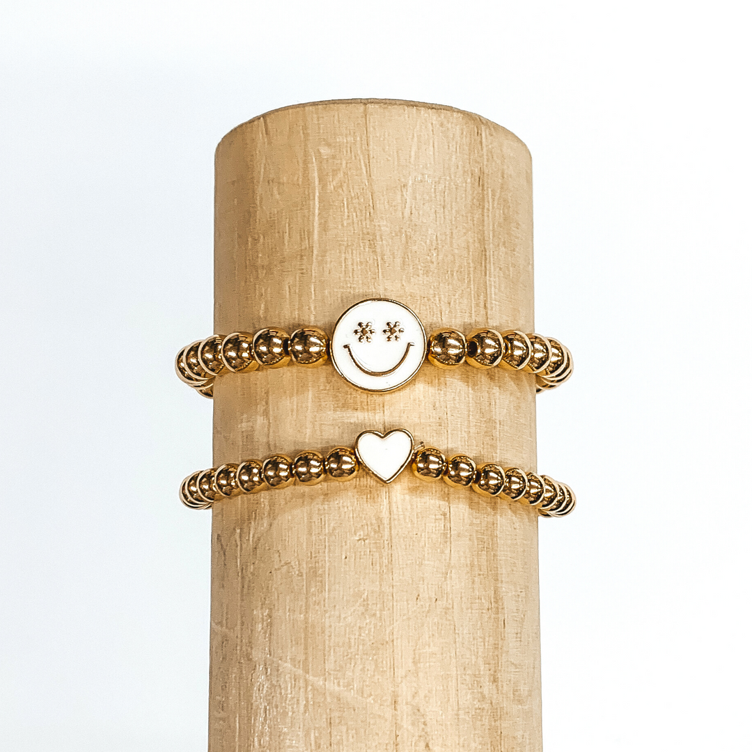 Two gold beaded bracelets. One bracelet has a tiny heart charm colored ivory. The other bracelet has a circle charm that is colored ivory with a smiley face in gold in the center. The eyes of the smiley face are tiny flowers. These two bracelets are pictured on a wood bracelet holder on a white background. 