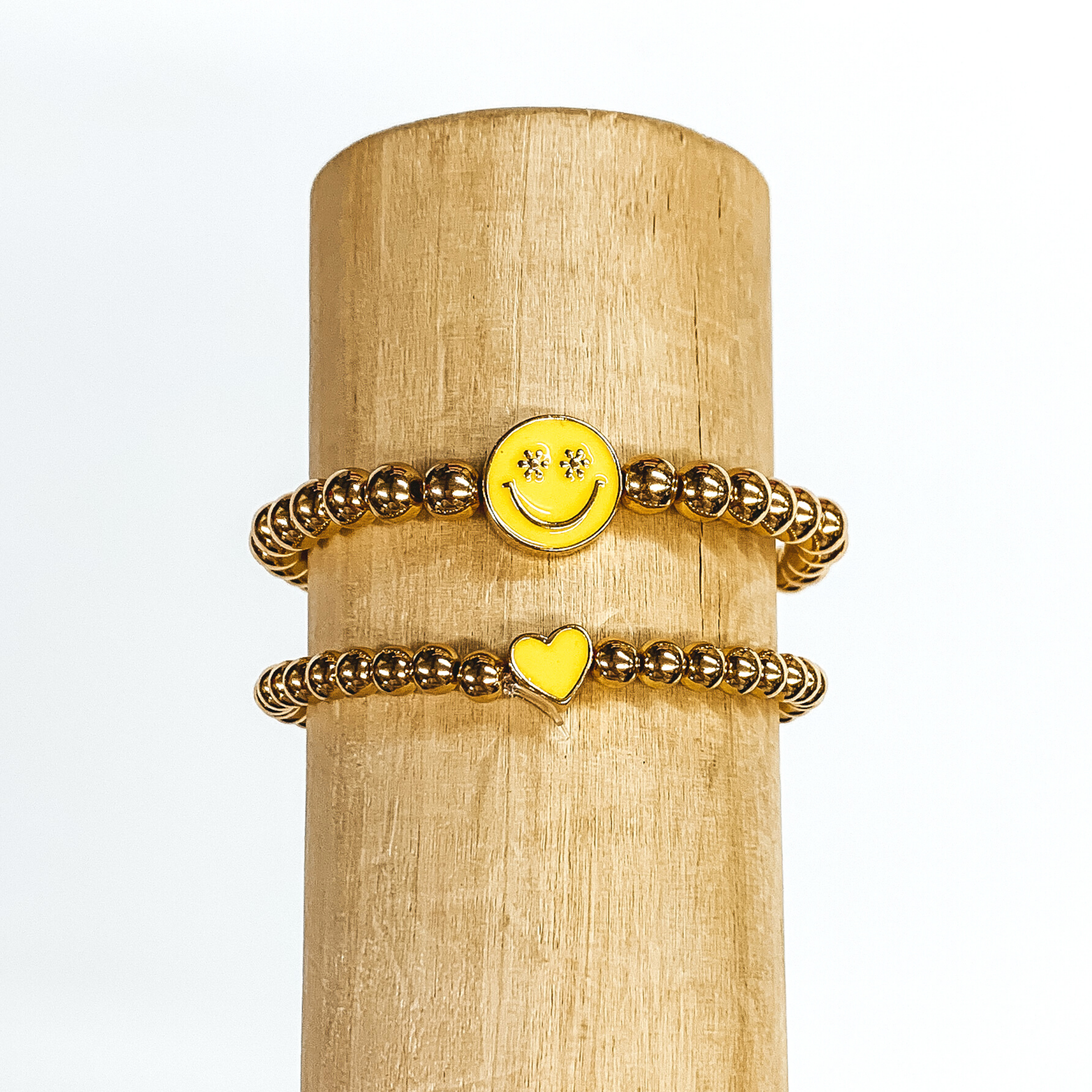 Two gold beaded bracelets. One bracelet has a tiny heart charm colored yellow. The other bracelet has a circle charm that is colored yellow with a smiley face in gold in the center. The eyes of the smiley face are tiny flowers. These two bracelets are pictured on a wood bracelet holder on a white background. 