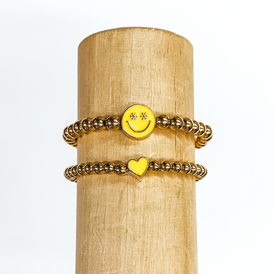 Two gold beaded bracelets. One bracelet has a tiny heart charm colored yellow. The other bracelet has a circle charm that is colored yellow with a smiley face in gold in the center. The eyes of the smiley face are tiny flowers. These two bracelets are pictured on a wood bracelet holder on a white background. 
