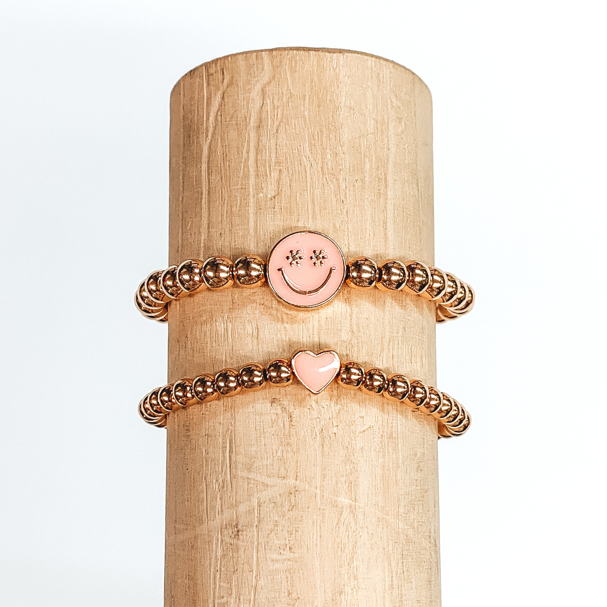 Two gold beaded bracelets. One bracelet has a tiny heart charm colored baby pink. The other bracelet has a circle charm that is colored baby pink with a smiley face in gold in the center. The eyes of the smiley face are tiny flowers. These two bracelets are pictured on a wood bracelet holder on a white background. 