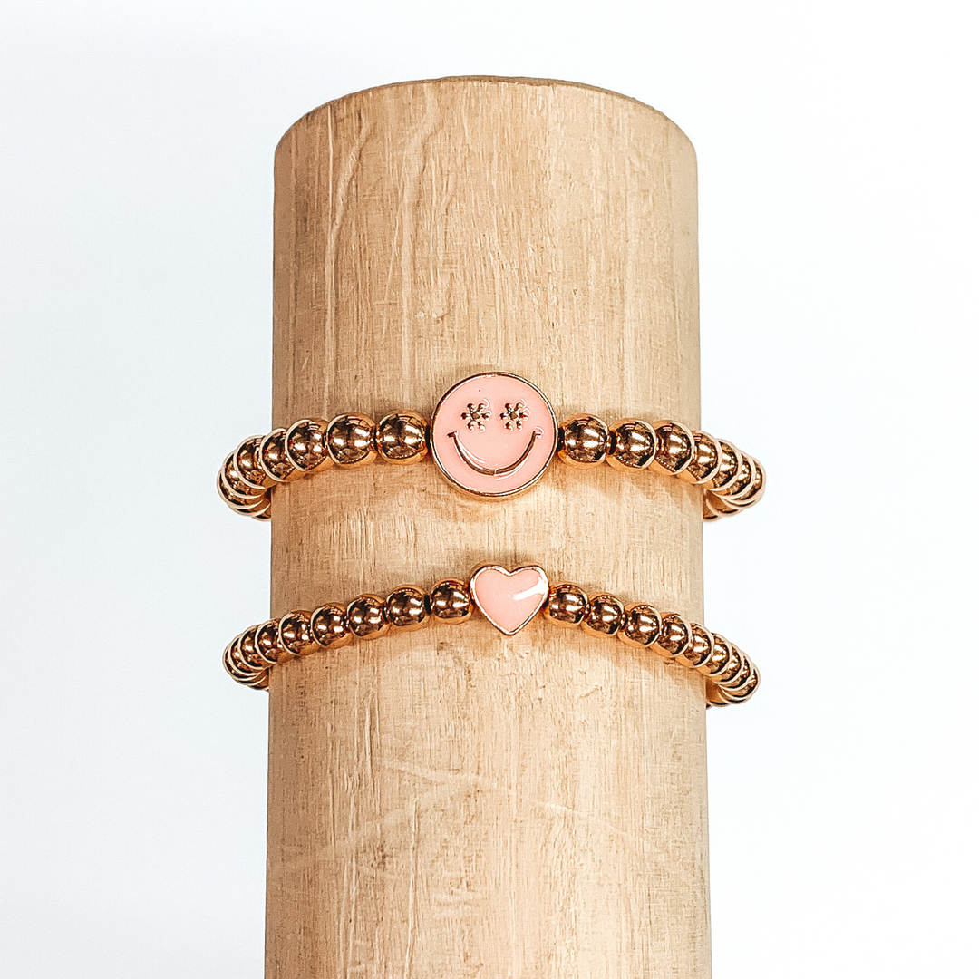 Two gold beaded bracelets. One bracelet has a tiny heart charm colored baby pink. The other bracelet has a circle charm that is colored baby pink with a smiley face in gold in the center. The eyes of the smiley face are tiny flowers. These two bracelets are pictured on a wood bracelet holder on a white background. 