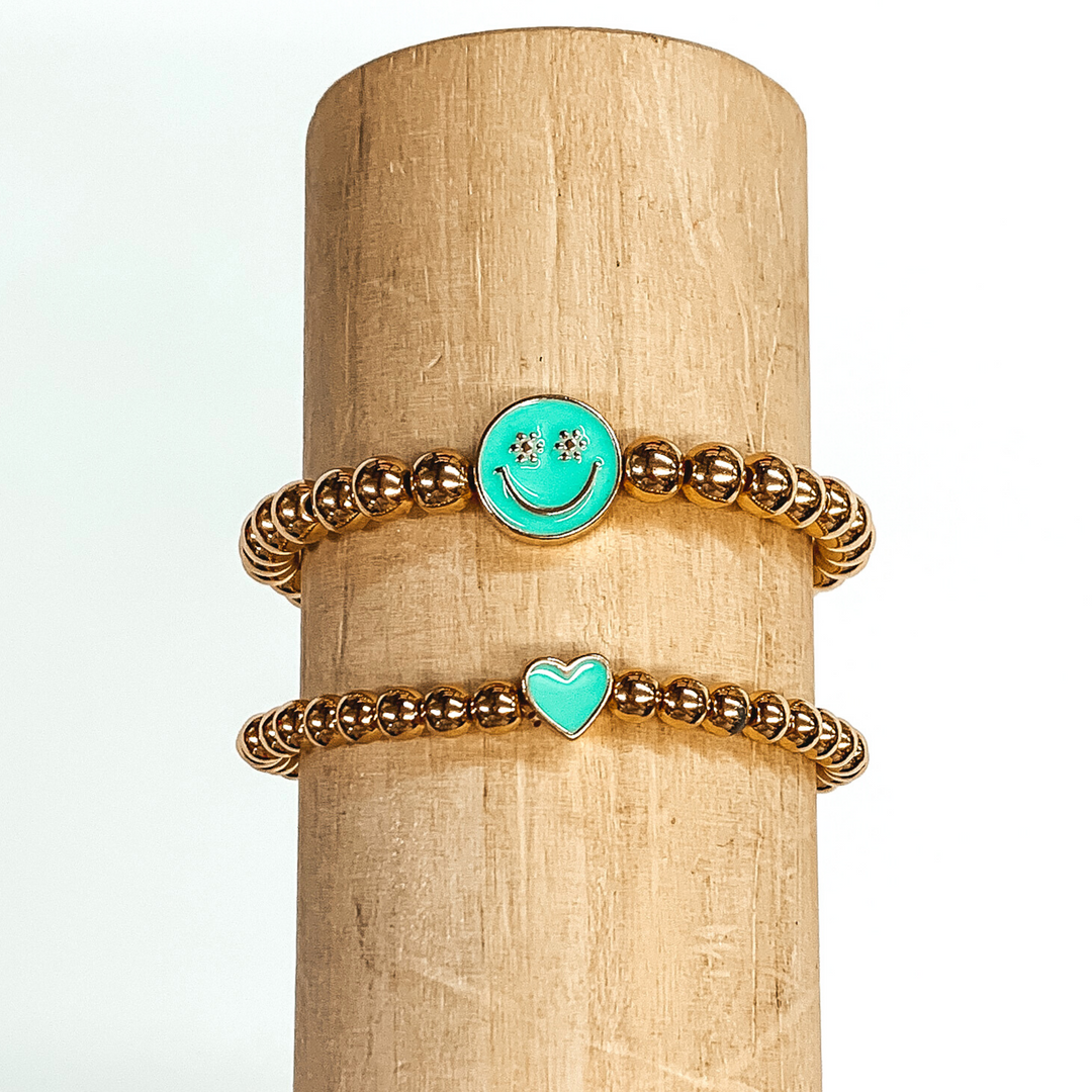 Two gold beaded bracelets. One bracelet has a tiny heart charm colored aqua. The other bracelet has a circle charm that is colored aqua with a smiley face in gold in the center. The eyes of the smiley face are tiny flowers. These two bracelets are pictured on a wood bracelet holder on a white background. 