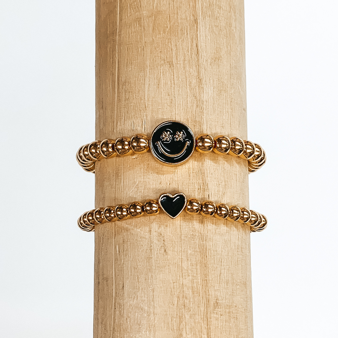 Two gold beaded bracelets. One bracelet has a tiny heart charm colored black. The other bracelet has a circle charm that is colored black with a smiley face in gold in the center. The eyes of the smiley face are tiny flowers. These two bracelets are pictured on a wood bracelet holder on a white background. 
