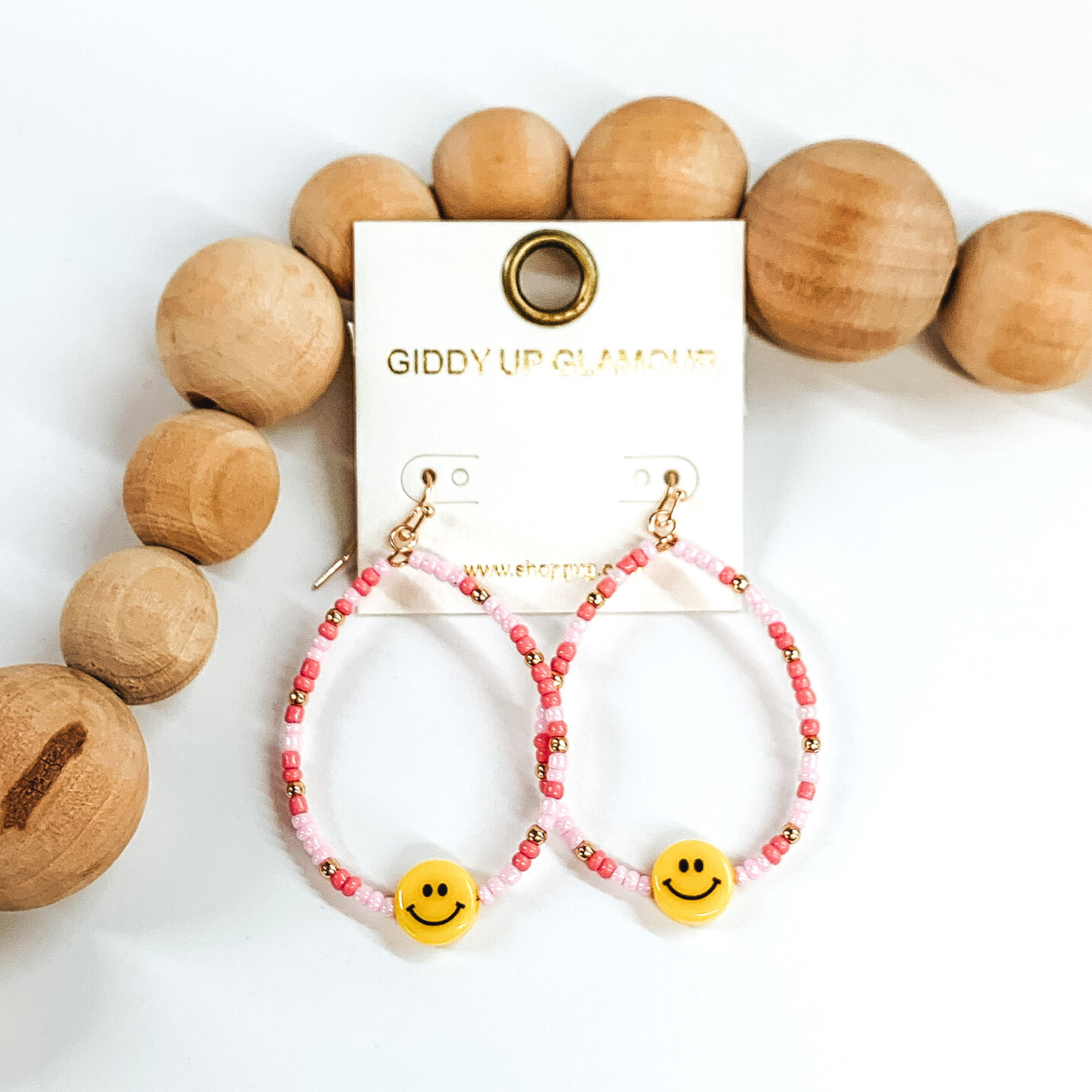 Hanging teardrop earrings with hanging yellow smiley faces. These earrings are covered in baby pink and pale pink beads with gold spacers. These earrings are pictured laying next to some tan beads on a white background.