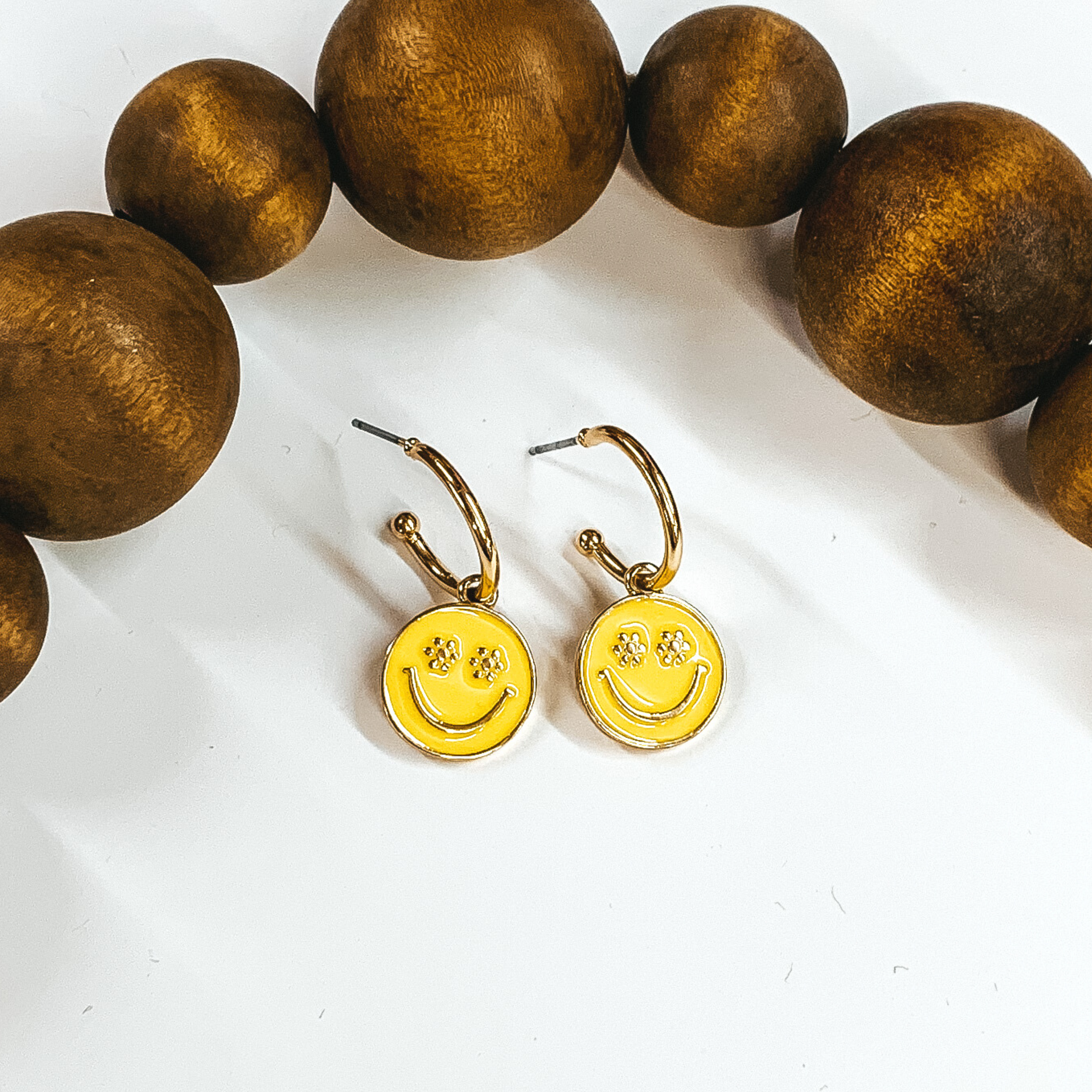 Small, gold hoop earrings with a hanging circle pendant in yellow. The pendant has a gold smiley face with the eyes being small flowers. These earrings are pictured laying on a white background with dark brown beads at the top of the picture. 