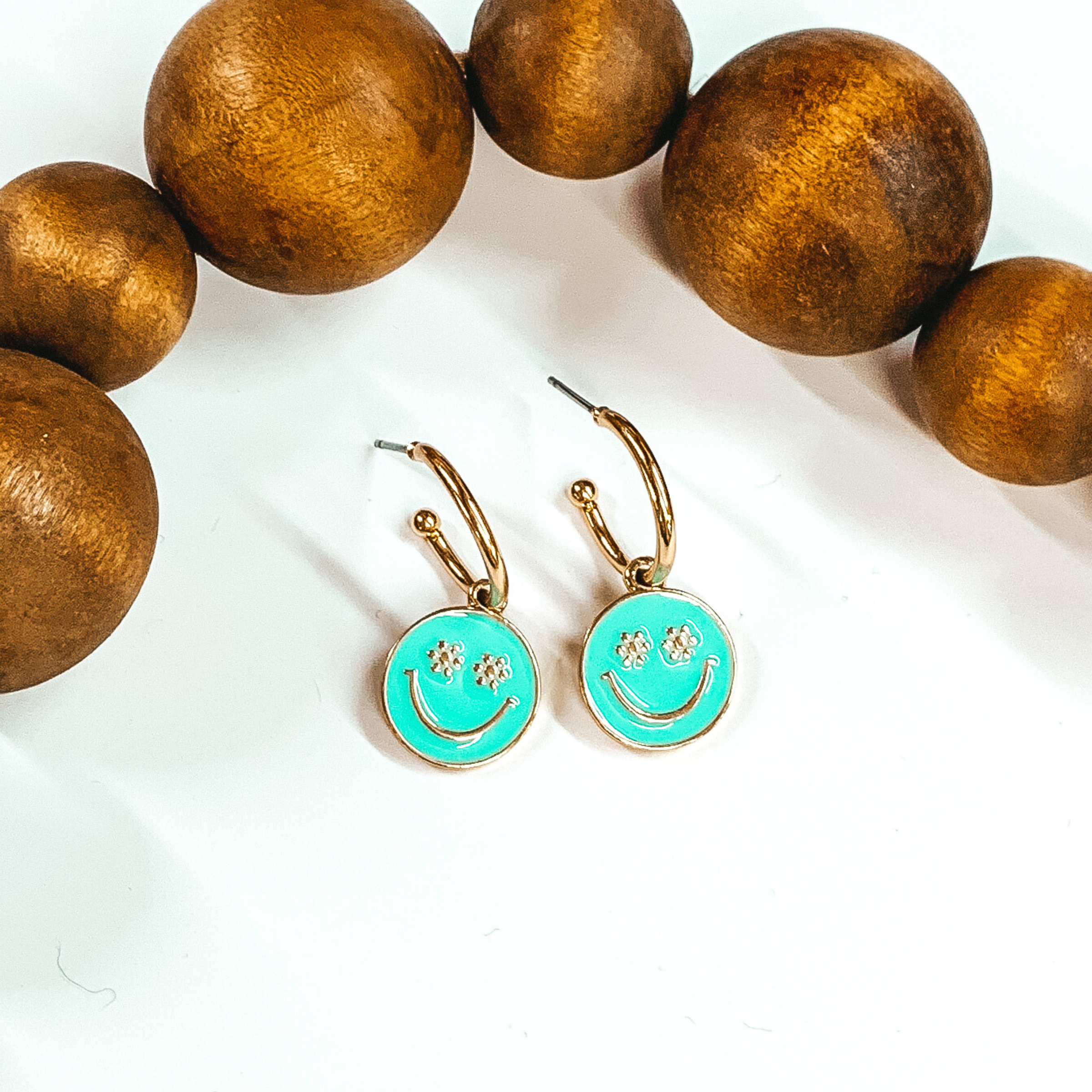 Small, gold hoop earrings with a hanging circle pendant in aqua. The pendant has a gold smiley face with the eyes being small flowers. These earrings are pictured laying on a white background with dark brown beads at the top of the picture. 