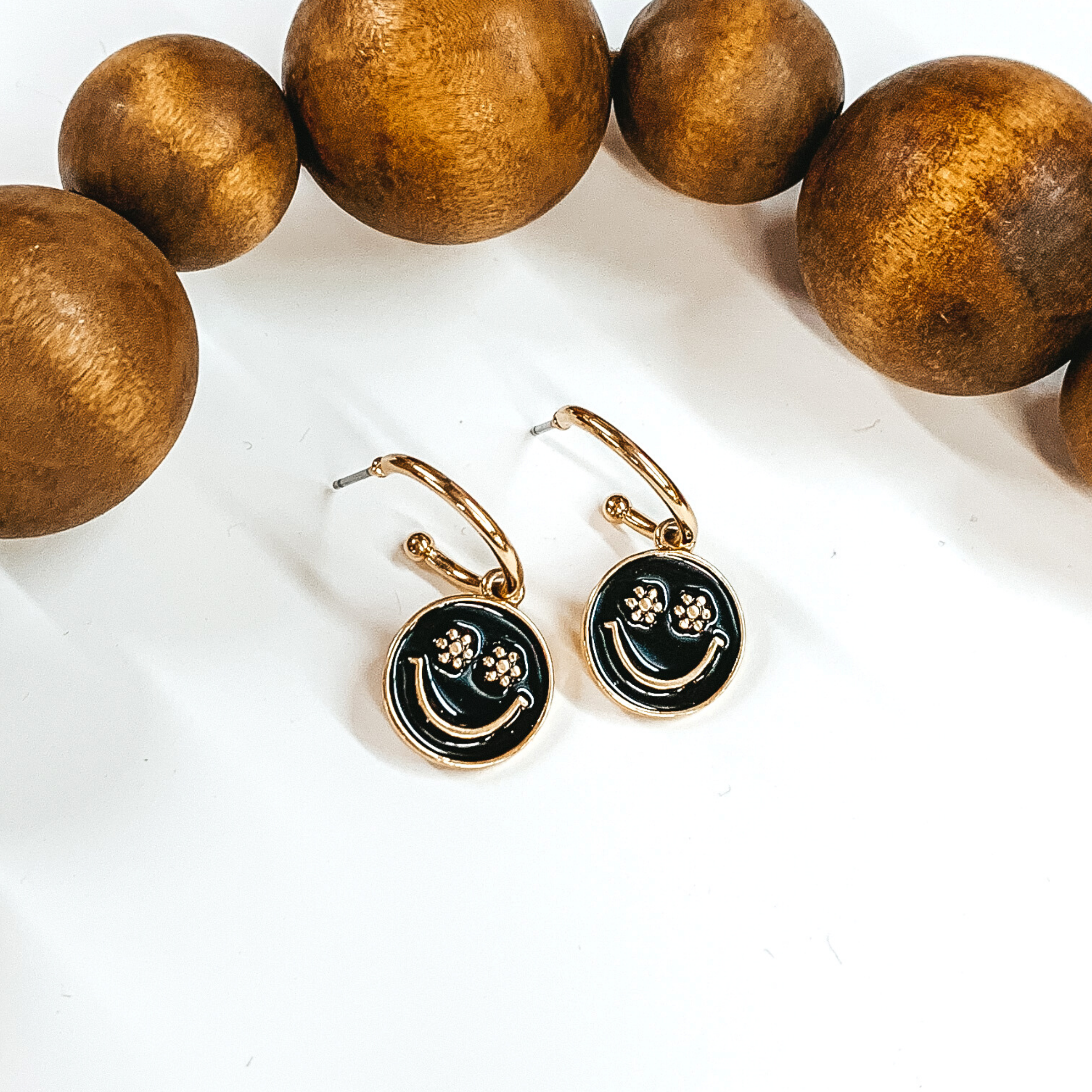 Small, gold hoop earrings with a hanging circle pendant in black. The pendant has a gold smiley face with the eyes being small flowers. These earrings are pictured laying on a white background with dark brown beads at the top of the picture. 