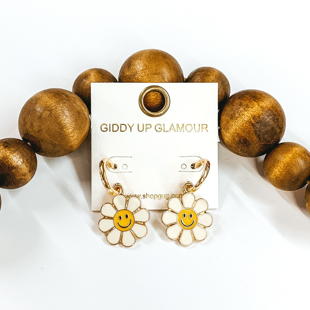 Small, gold hoop earrings with a hanging flower pendant in ivory with a yellow center. The pendant has a black smiley face in the center of the flower. These earrings are pictured laying on a white background against dark brown beads towards the top of the picture. 