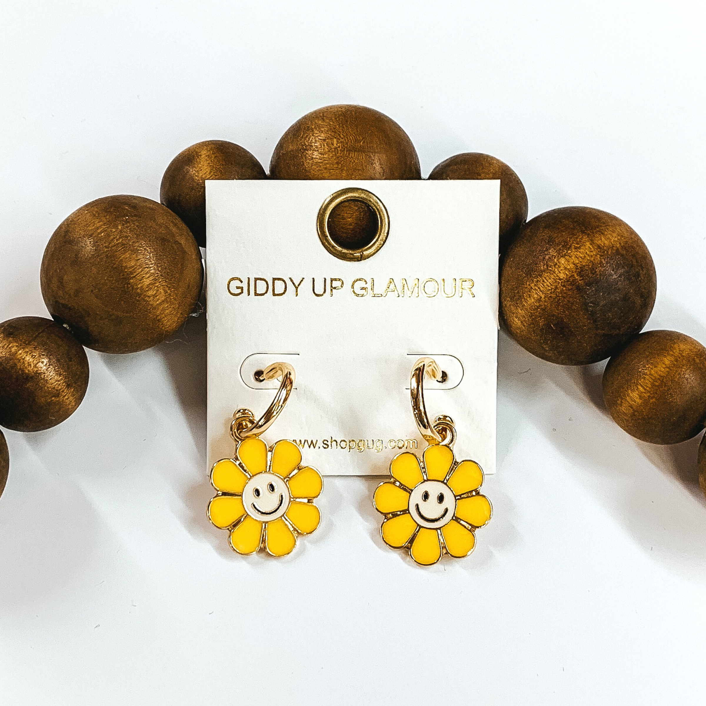 Small, gold hoop earrings with a hanging flower pendant in yellow with an ivory center. The pendant has a black smiley face in the center of the flower. These earrings are pictured laying on a white background against dark brown beads towards the top of the picture. 