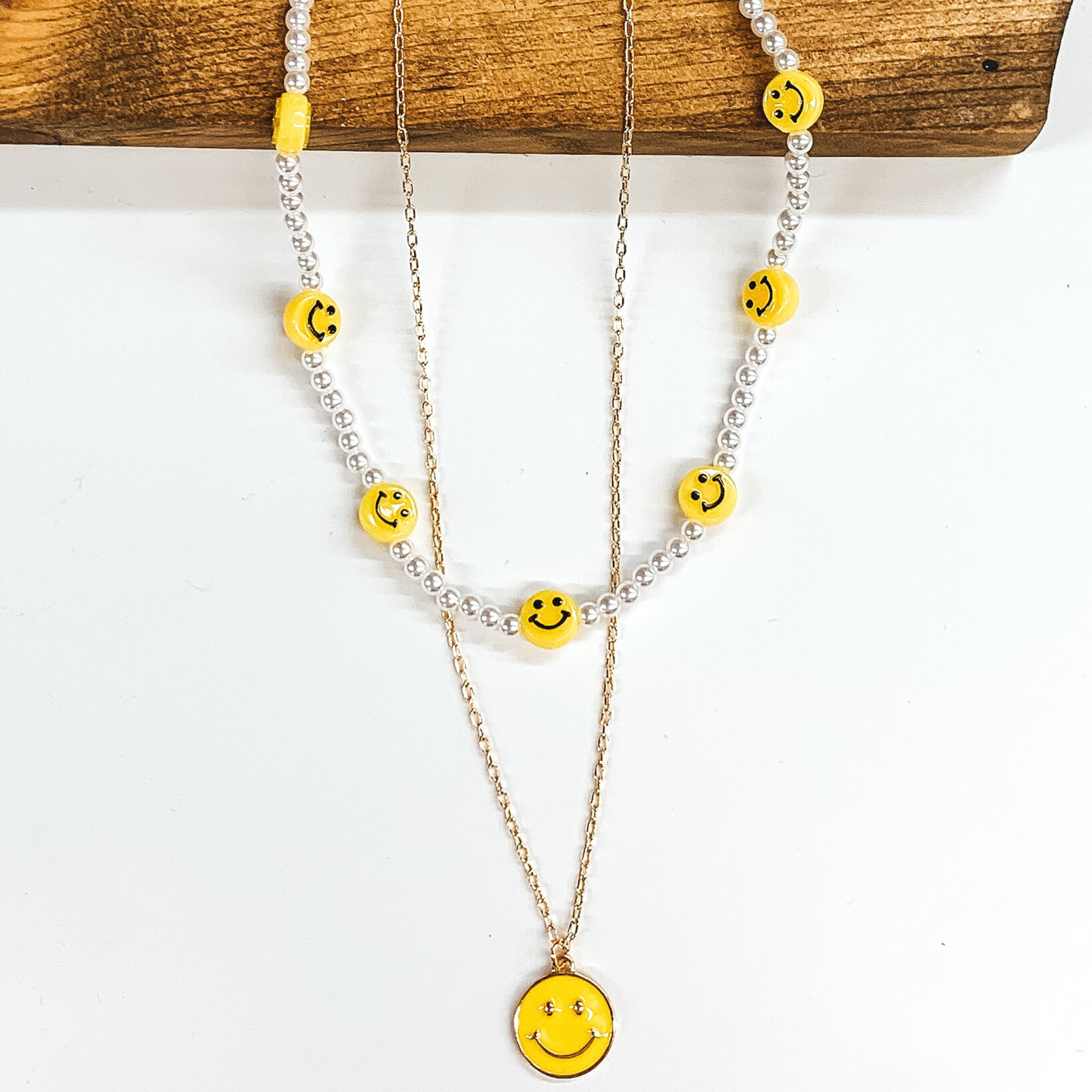 Double layered necklace. The white beaded strand has yellow smiley face charm spacers. The other strand is a gold chained necklace with a yellow circle pendant with a gold smiley face. This necklace is pictures partially laying on a dark piece of wood on a white background. 