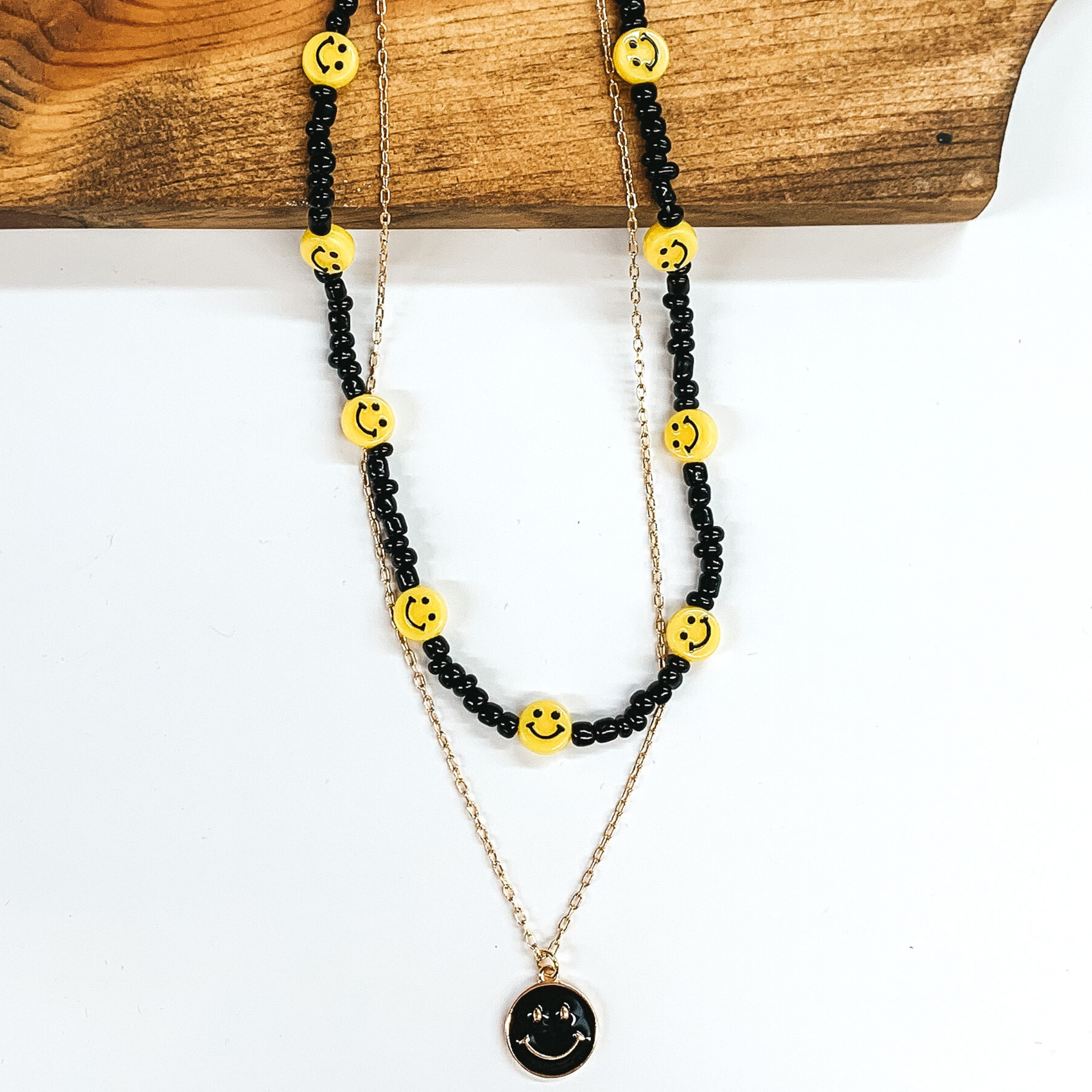Double layered necklace. The black beaded strand has yellow smiley face charm spacers. The other strand is a gold chained necklace with a yellow circle pendant with a gold smiley face. This necklace is pictures partially laying on a dark piece of wood on a white background.