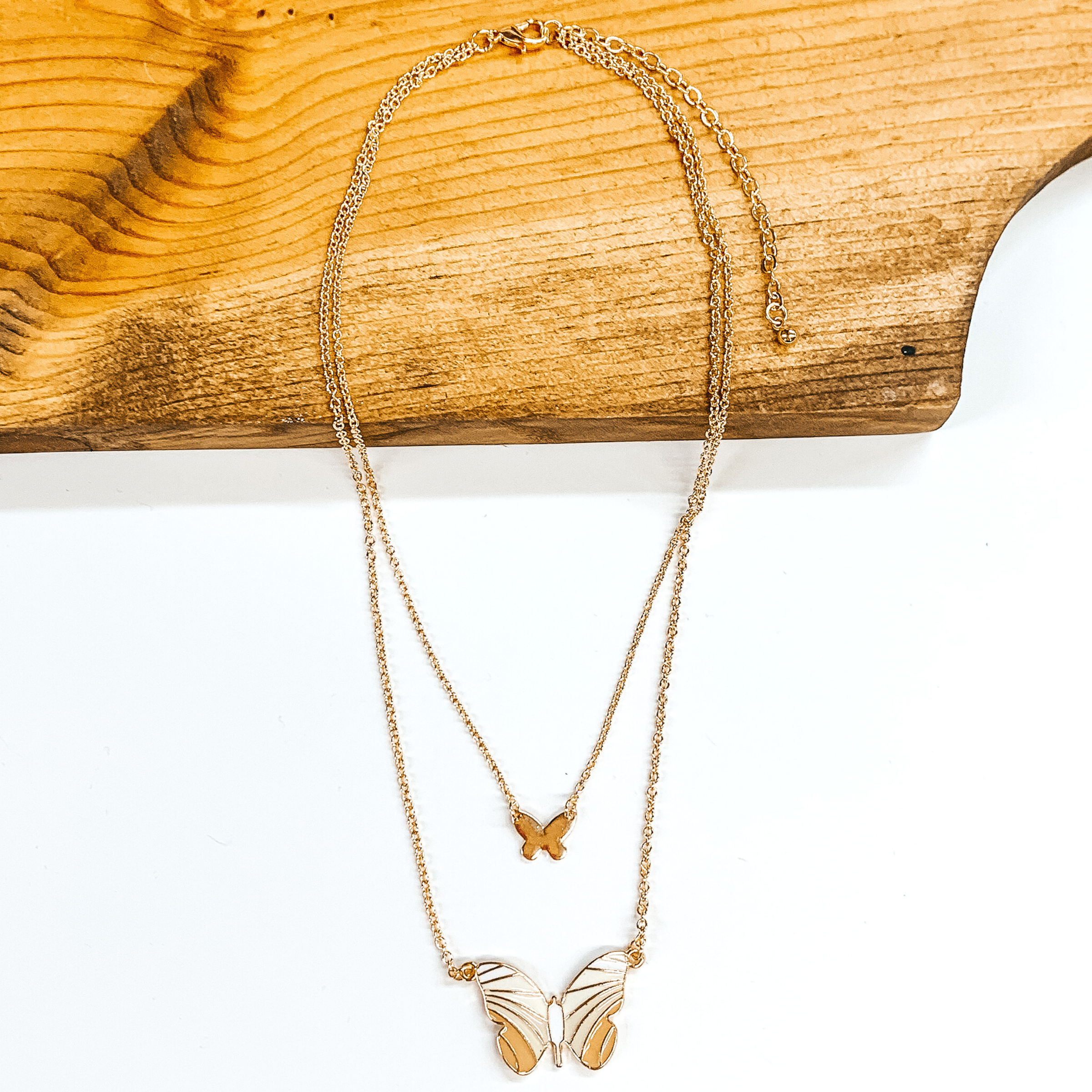 This a double gold chained necklace. The shorter chain has a small gold butterfly chain. The longer strand has a bigger butterfly pendant. The colored of the wings have colors starting off with white and getting darker to beige at the bottom of the wings. This necklace is partially laying on a dark piece of wood on a white background. 