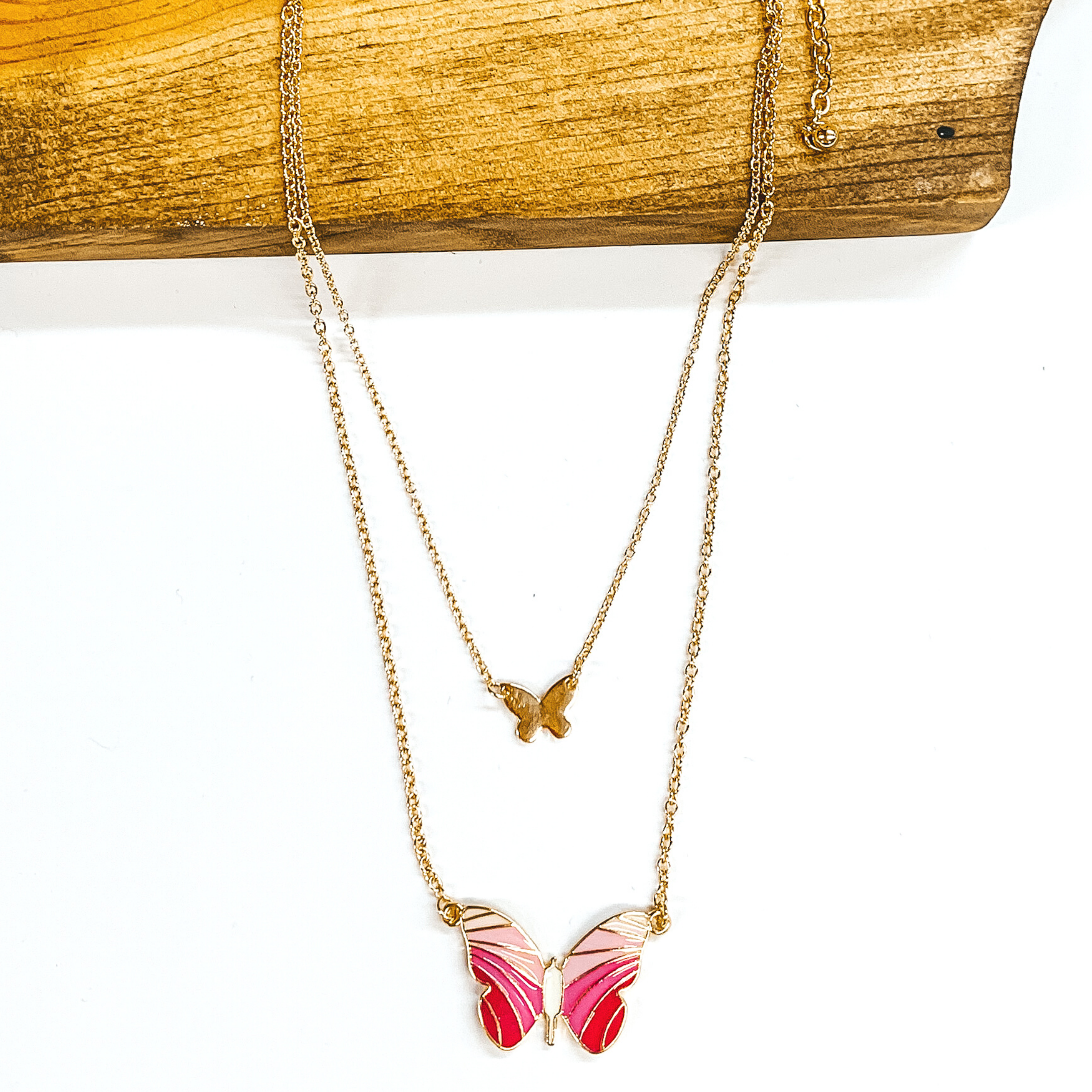 This a double gold chained necklace. The shorter chain has a small gold butterfly chain. The longer strand has a bigger butterfly pendant. The colored of the wings have colors starting off with the lightest pink and getting darker to a darker pink at the bottom of the wings. This necklace is partially laying on a dark piece of wood on a white background. 