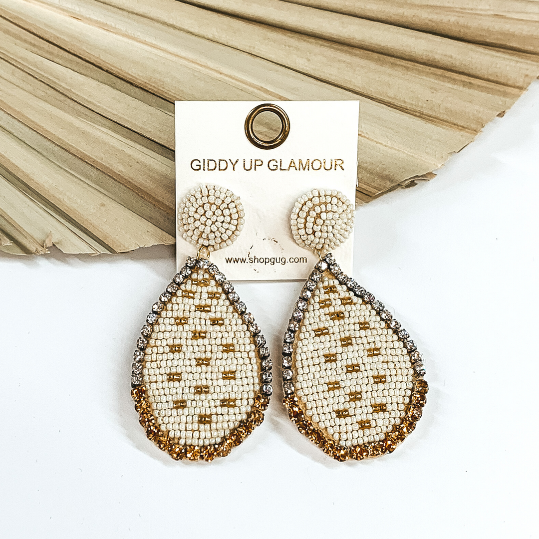 These earrings have a beaded circle stud with a hanging beaded teardrop pendant. The pendant is covered with ivory beads with gold beads spread throughout evenly. The teardrop pendant is outlined hald with clear crystals and half in gold crystals. These earrings are pictured in ront of a pale green leaf and on a white background. 