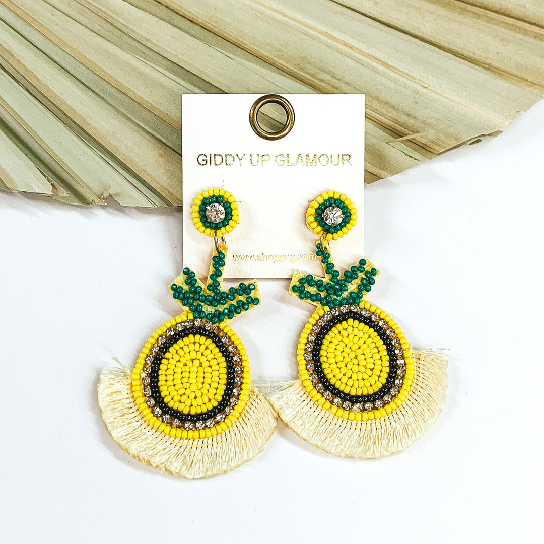 These earrings have circle stud earrings with hanging pineapple shaped pendants with light yellow fringe on the bottom of the earrings. These earrings include yellow, green, and black beads. There are also clear crystals. These earrings are pictured laying in front of a pale green leaf and on a white background.