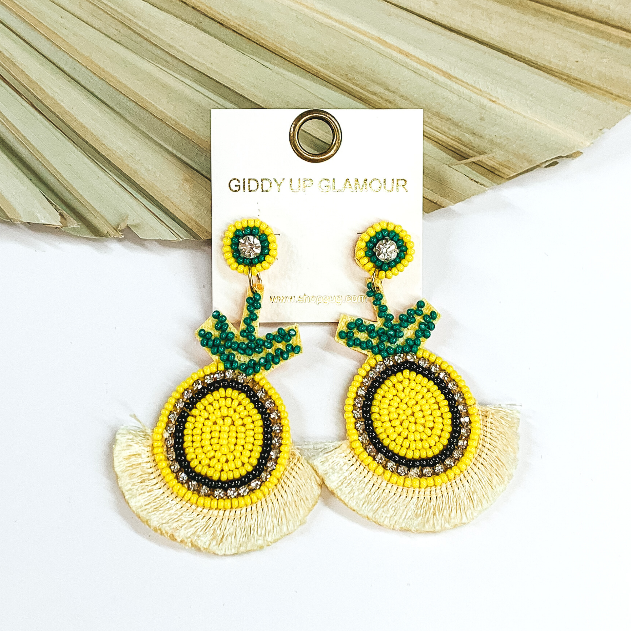 These earrings have circle stud earrings with hanging pineapple shaped pendants with light yellow fringe on the bottom of the earrings. These earrings include yellow, green, and black beads. There are also clear crystals. These earrings are pictured laying in front of a pale green leaf and on a white background.