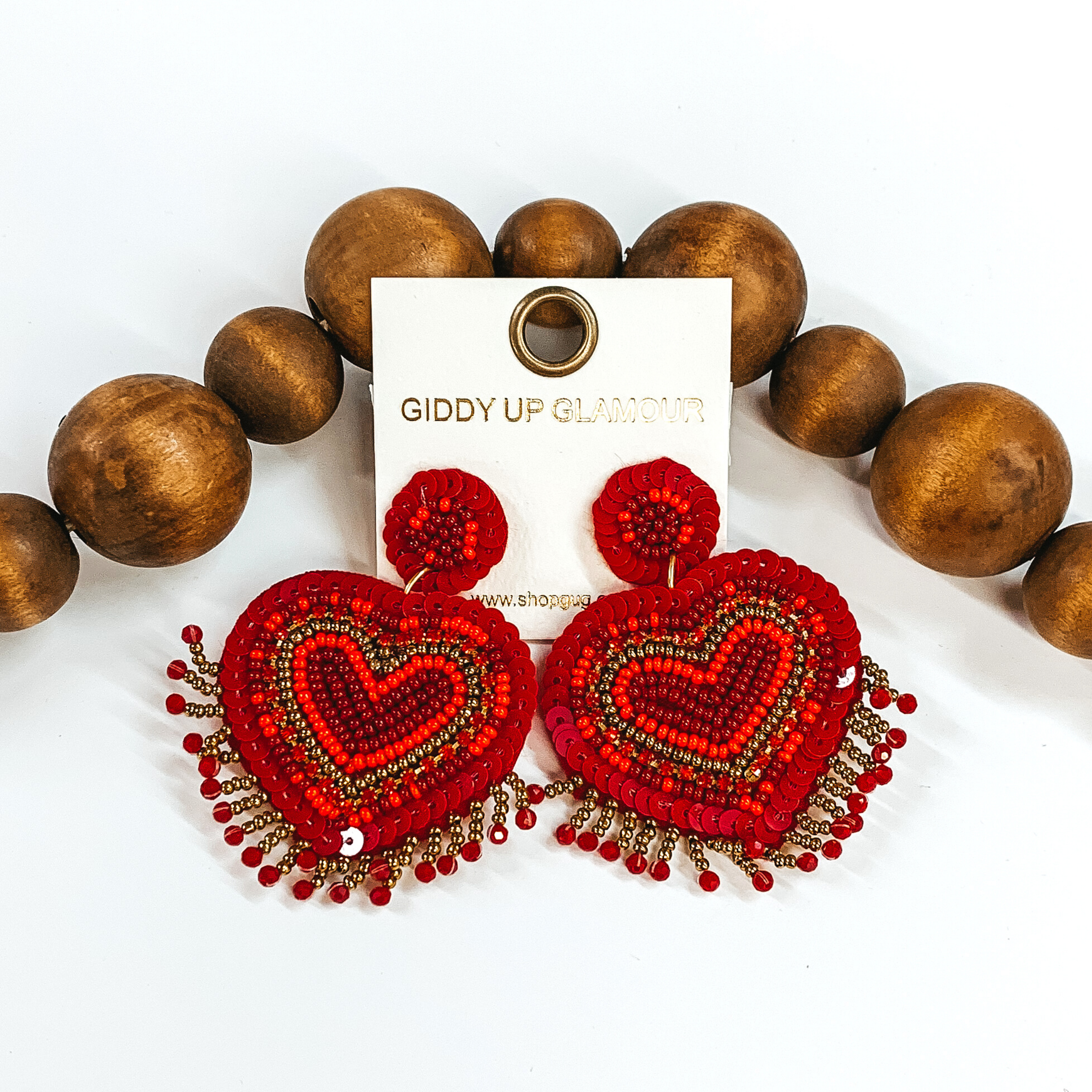 Sequin and beaded heart and dangle earrings with gold beaded fringe. It includes red, dark red, and gold beads and light pink sequins. These earrings are laying in front of dark brown beads on a white background.  