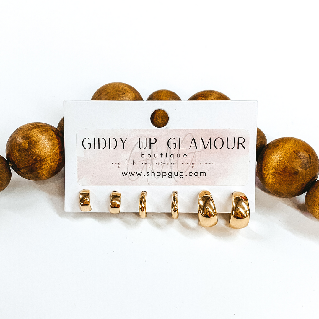 This is a 3 piece gold rounded hoop set. These are small hoops included one half hoop set and two full hoop sets These earrings are pictured on a Giddy Up Glamour earring holder laying in front of dark brown beads on a white background.  
