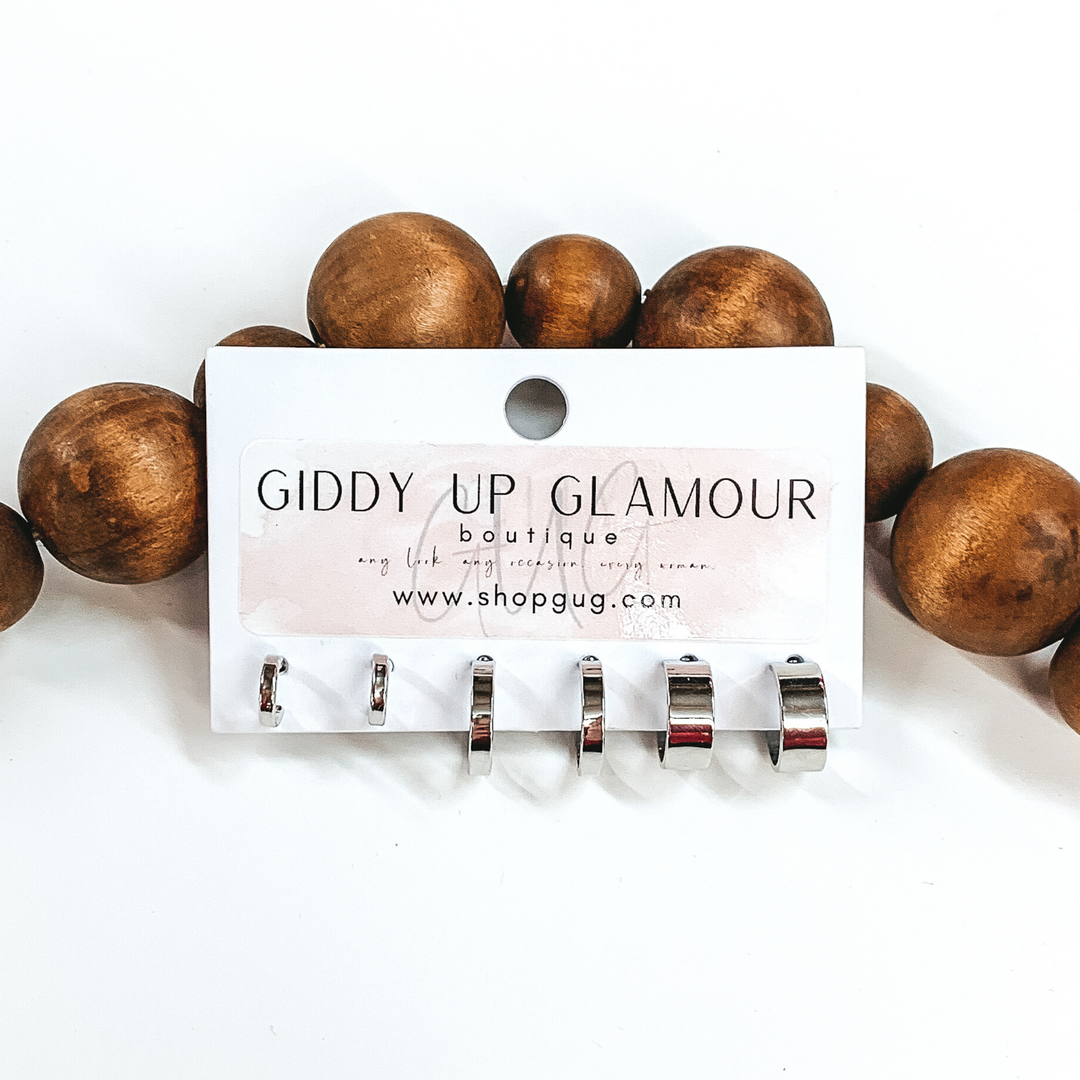 This is a 3 piece silver flat hoop set. All three hoops have different widths. These earrings are pictured on a Giddy Up Glamour earring holder laying in front of dark brown beads on a white background.